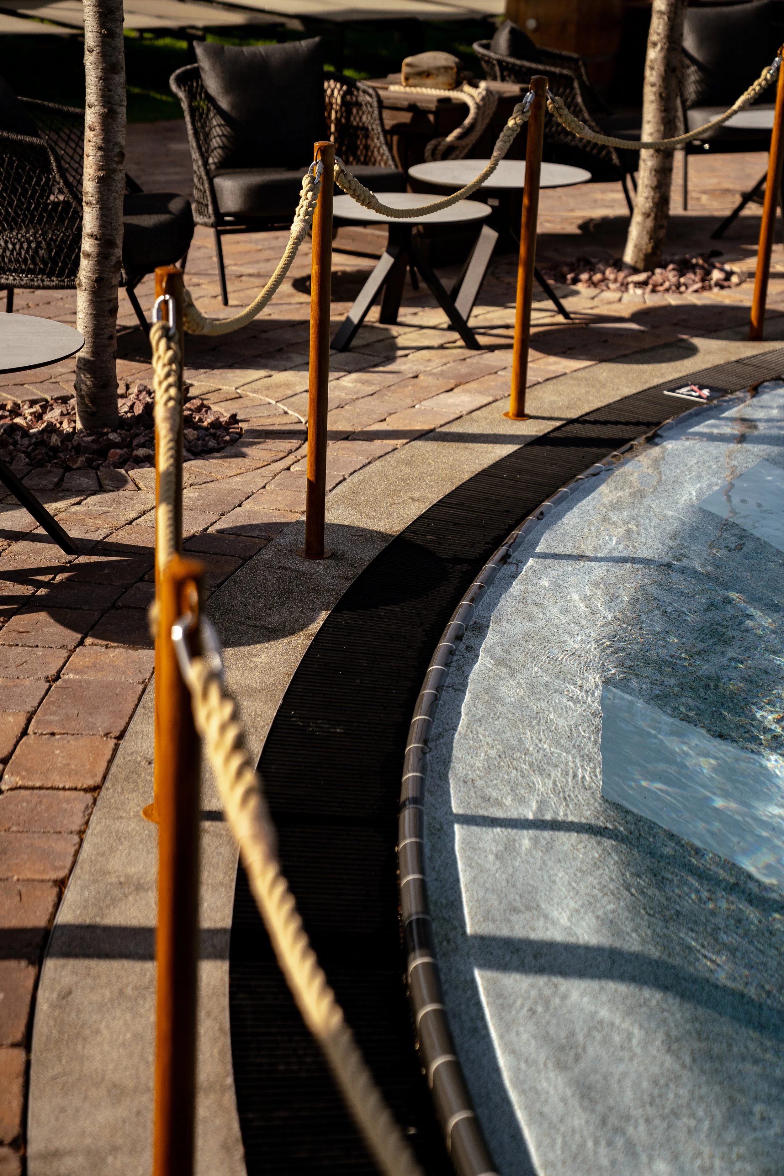 Sitzgelegenheiten mit schwarzen Stühlen und Tischen, umgeben von Bäumen und eine Absperrung entlang des Schwimmbads.