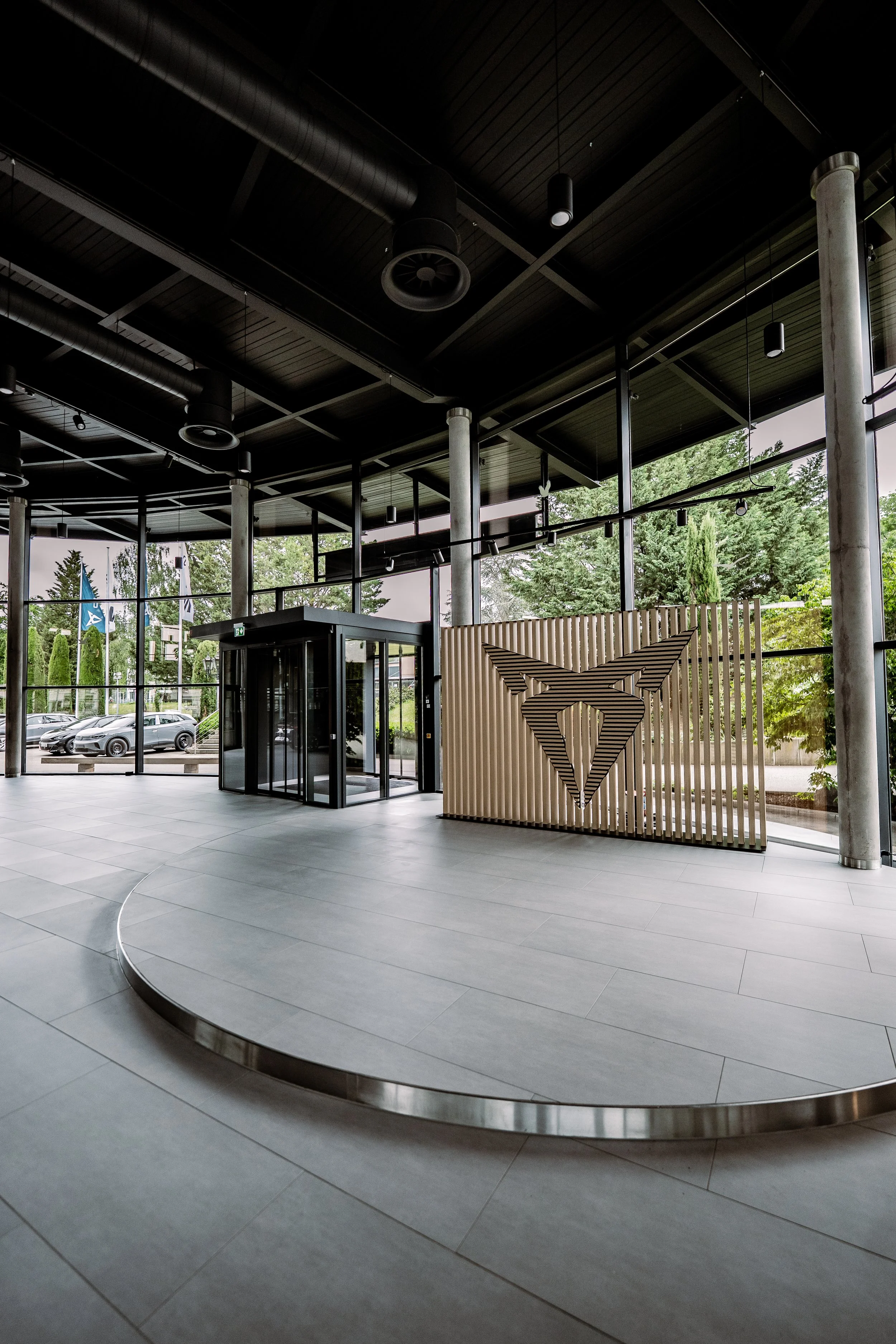 Ein moderner Eingangsbereich mit futuristischem Design, große Glasfenster, ein Empfangstresen, dekoratives Element mit einem abstrakten Logo aus Holzlamellen und ein Drehkreuz für Zugangskontrolle.