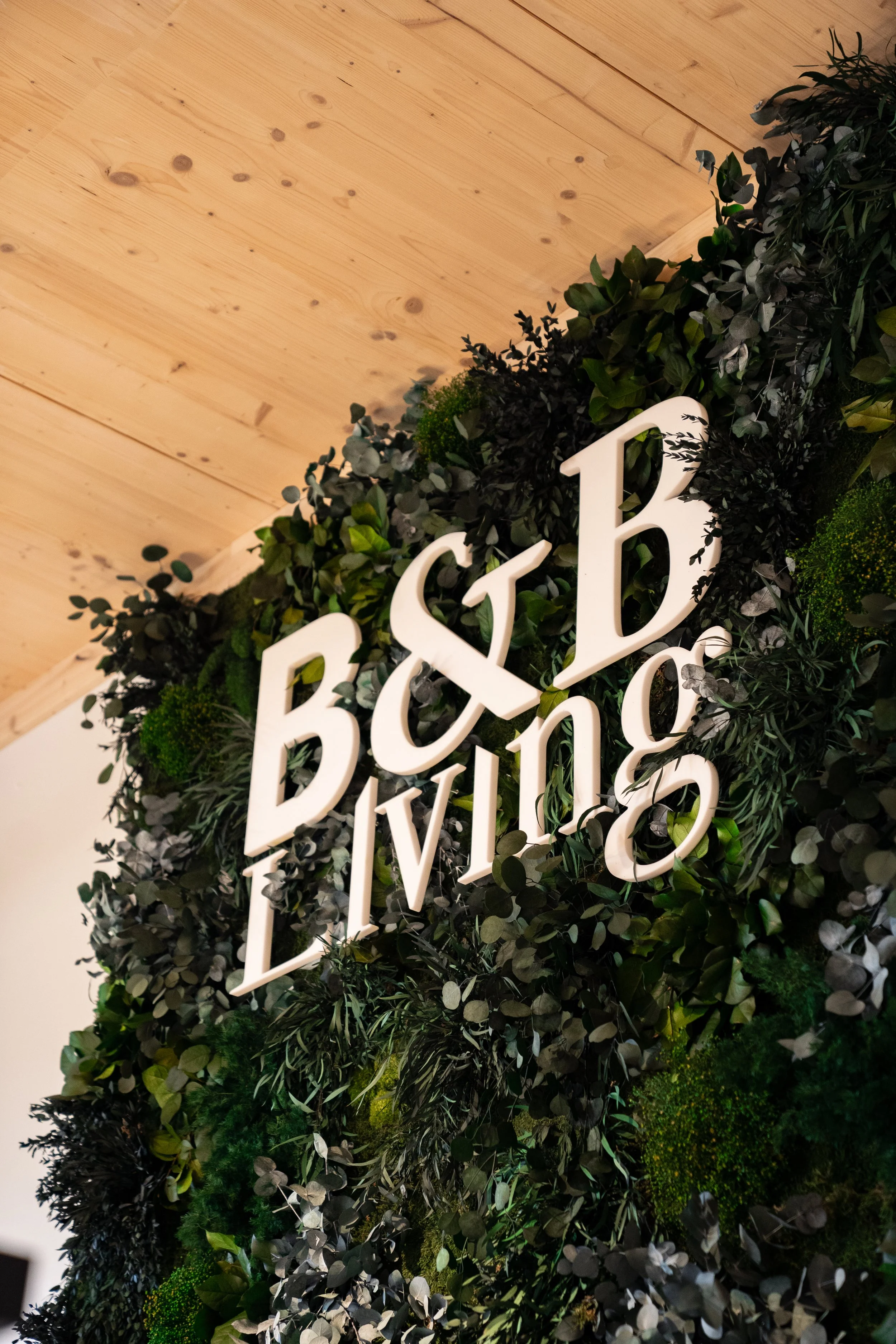 Grüner Wanddekoration mit weißen Buchstaben, die 'B&B' und 'LIVING' lesen, umgeben von dunklen und grünen Blättern, an einer Holzdecke.
