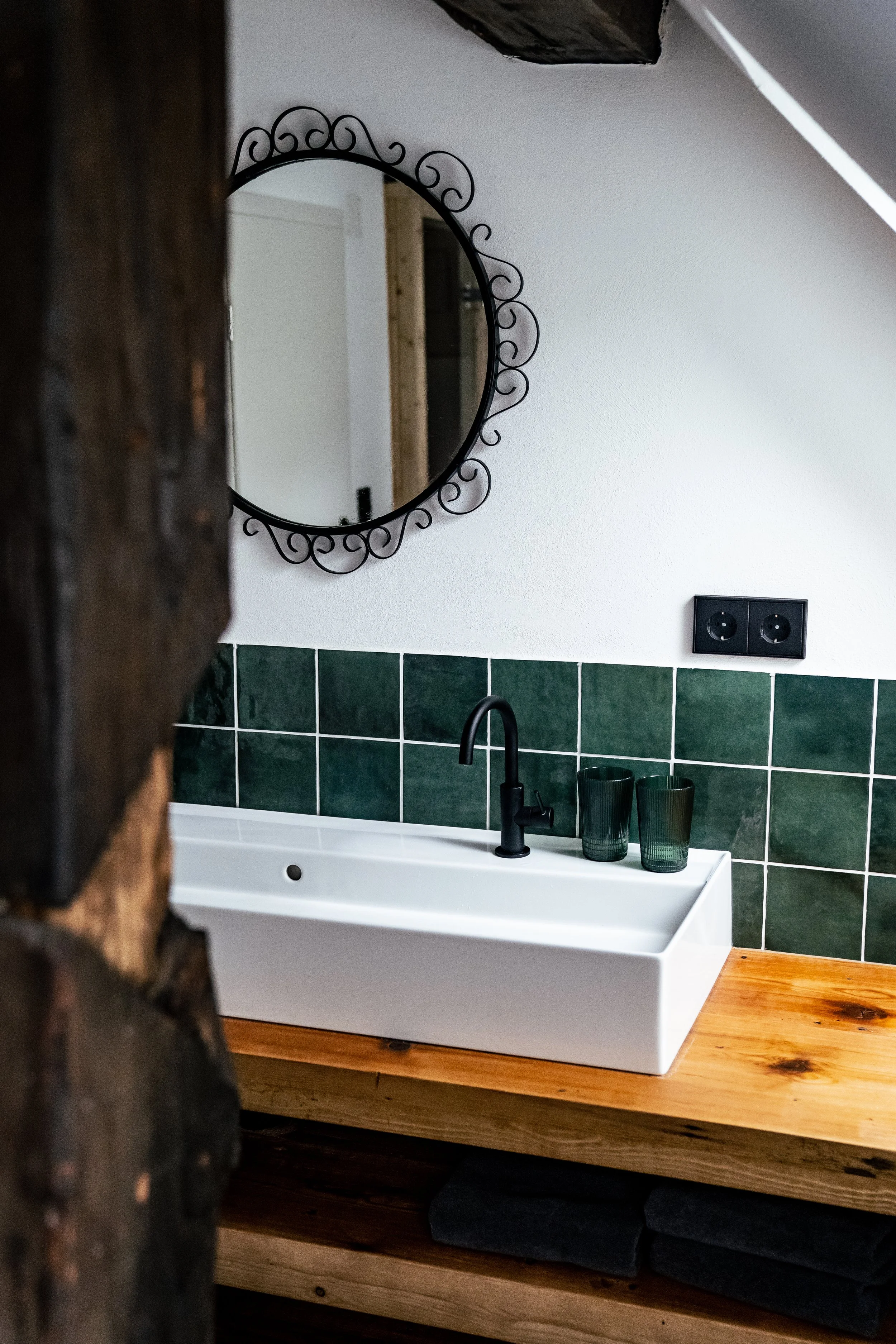 Schmales Badezimmer mit Wandspiegel, grünen Fliesen, weißem Waschbecken, schwarzer Armatur und zwei grünen Gläsern auf Holzarbeitsplatte.
