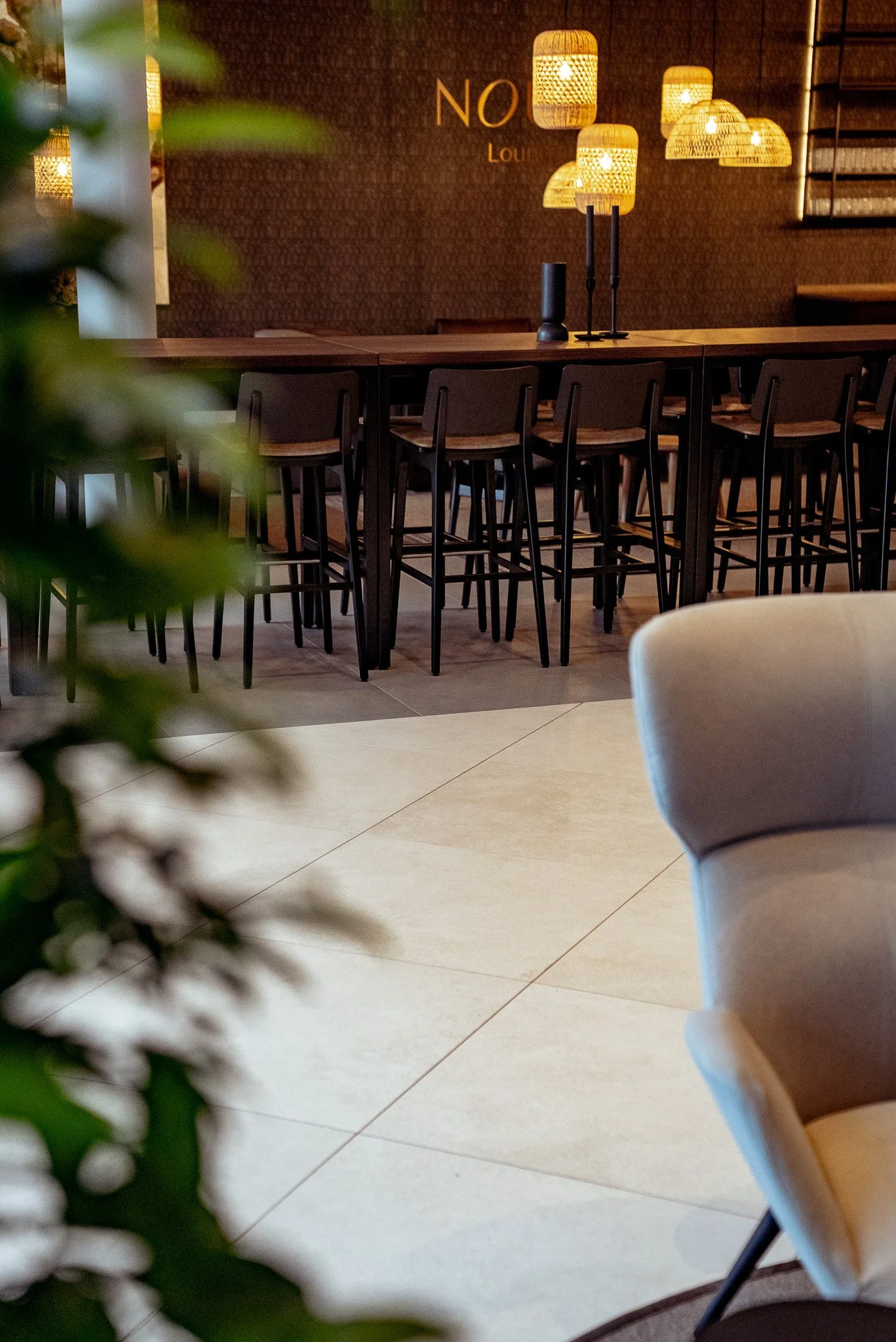 Ein moderner Lounge-Bereich mit hohen schwarzen Stühlen an einem langen Holztresen, der mit Tischlampen und Dekoration beleuchtet ist. Im Hintergrund die Wand mit dem Wort 'NO' und später 'Lounge'. Im Vordergrund eine graue Sitzecke und eine grüne Pf