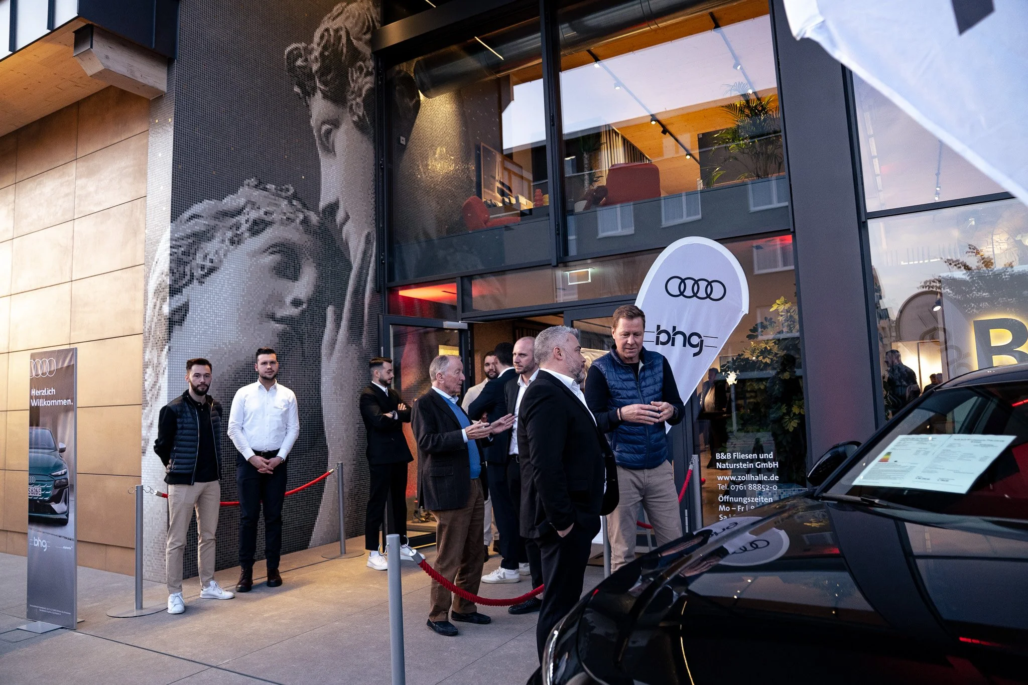 Menschen stehen vor einem Ladengeschäft mit einem großen Mosaikbild eines Frauengesichts und einem Aufsteller mit Audiall-Logo. Es gibt eine Warteschlange, rotes Absperrband und ein schwarzes Auto vorne.