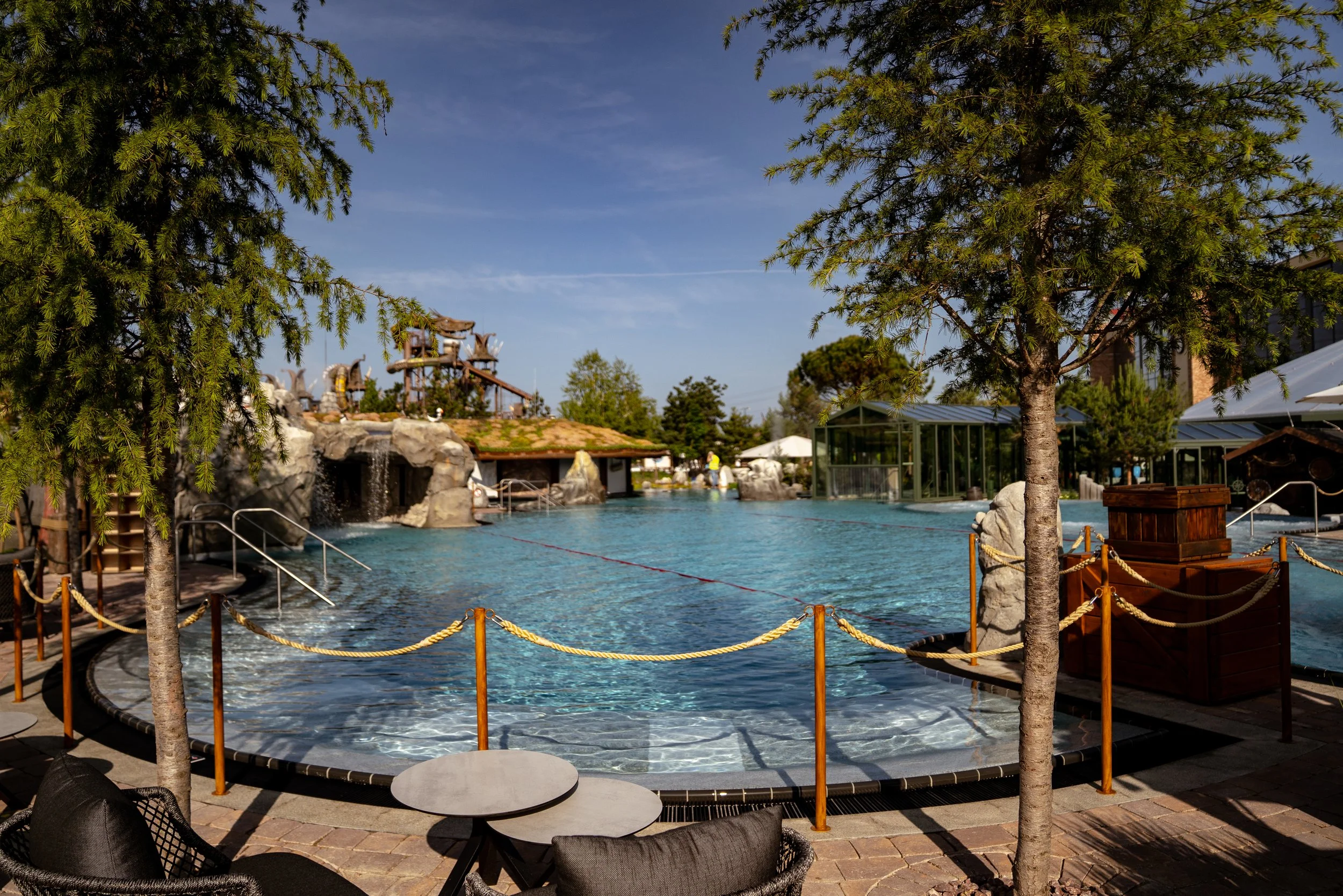 Ein großer, gepflegter Außenpool mit Sonnenliegen, umgeben von Bäumen und einem Zaun, bei sonnigem Himmel, im Hintergrund befinden sich Wasserattraktionen und ein modernes Gebäudekomplex.
