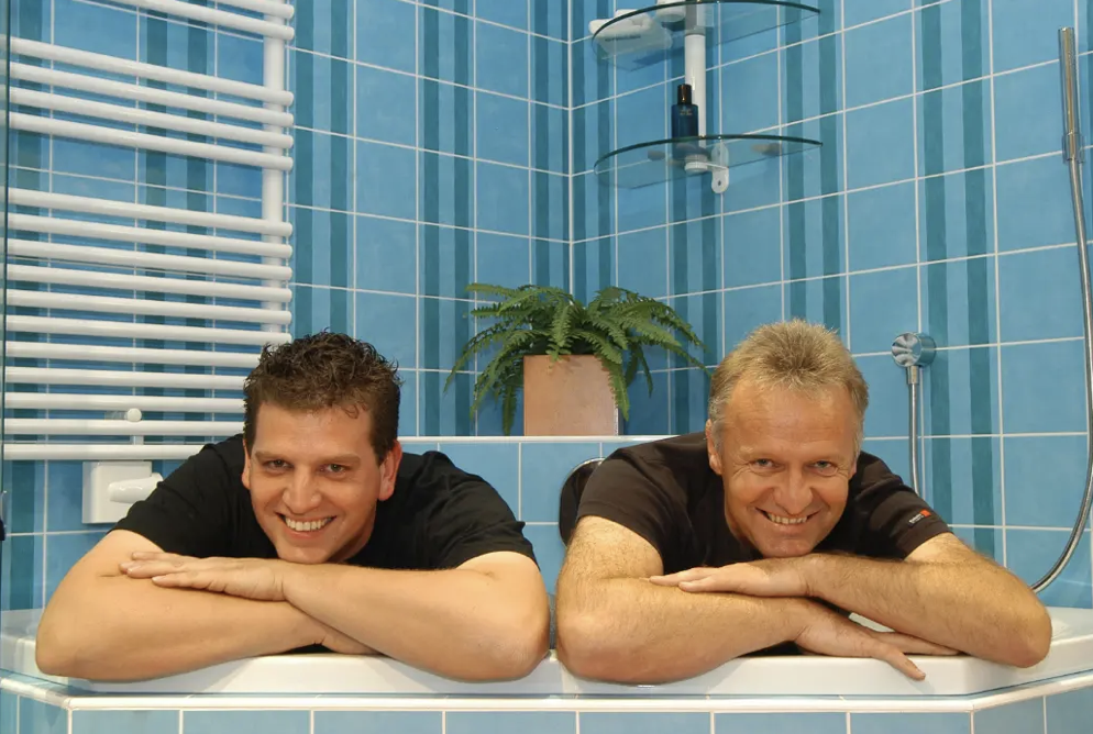 Zwei Männer liegen entspannt in einer Badewanne im Badezimmer, lächelnd zur Kamera schauend.