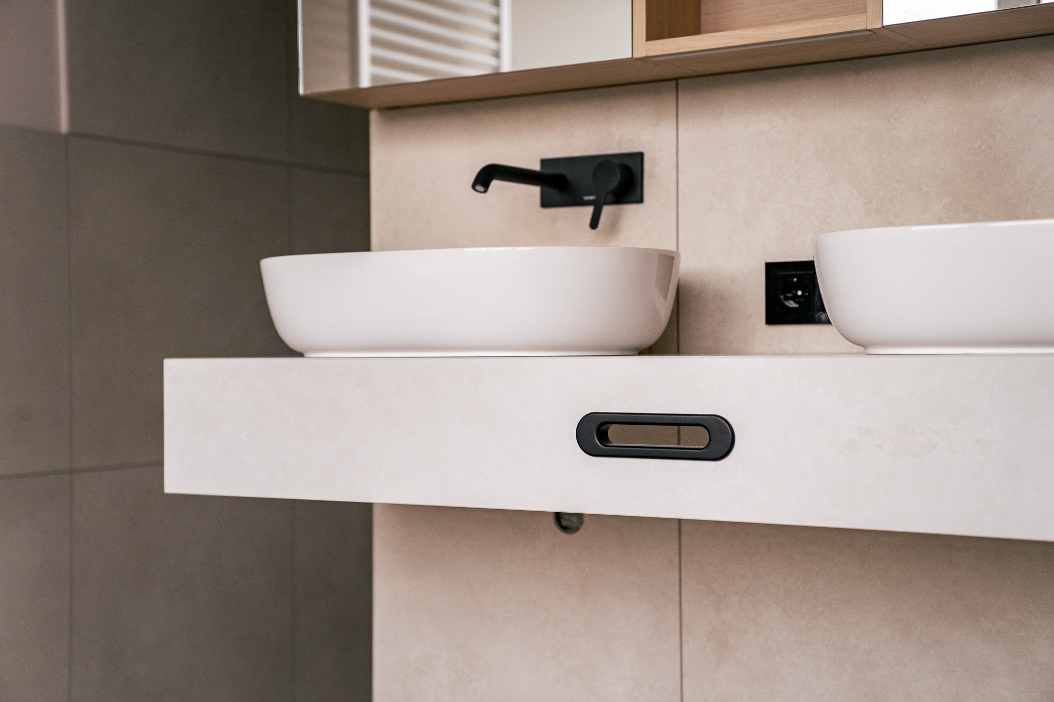 Weiße Waschbecken auf einem weißen Waschtisch in einem Badezimmer, schwarzer Wasserhahn und Steckdose auf beige Wandfliesen.