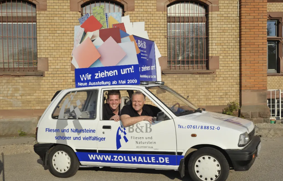 Zwei Männer sitzen in einem weißen kleinen Auto mit Werbung für Fliesen und Natursteine von B&B, vor einem Backsteingebäude, mit einem großen umziehenden Symbol auf dem Dach, das auf eine Ausstellung im Mai 2009 hinweist.