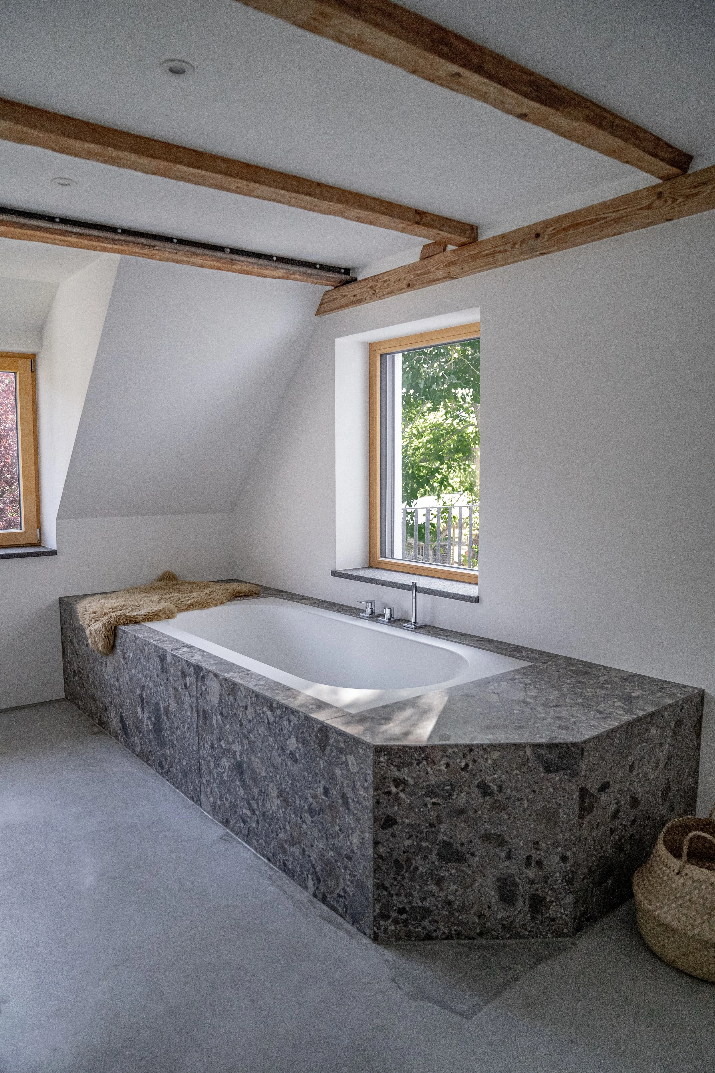 Ein Badezimmer mit einer freistehenden Badewanne, umgeben von einer Steinverkleidung, vor einer Fensterfront mit Blick ins Grüne. Das Zimmer hat einen schrägen Deckenabschnitt mit sichtbaren Holzbalken.