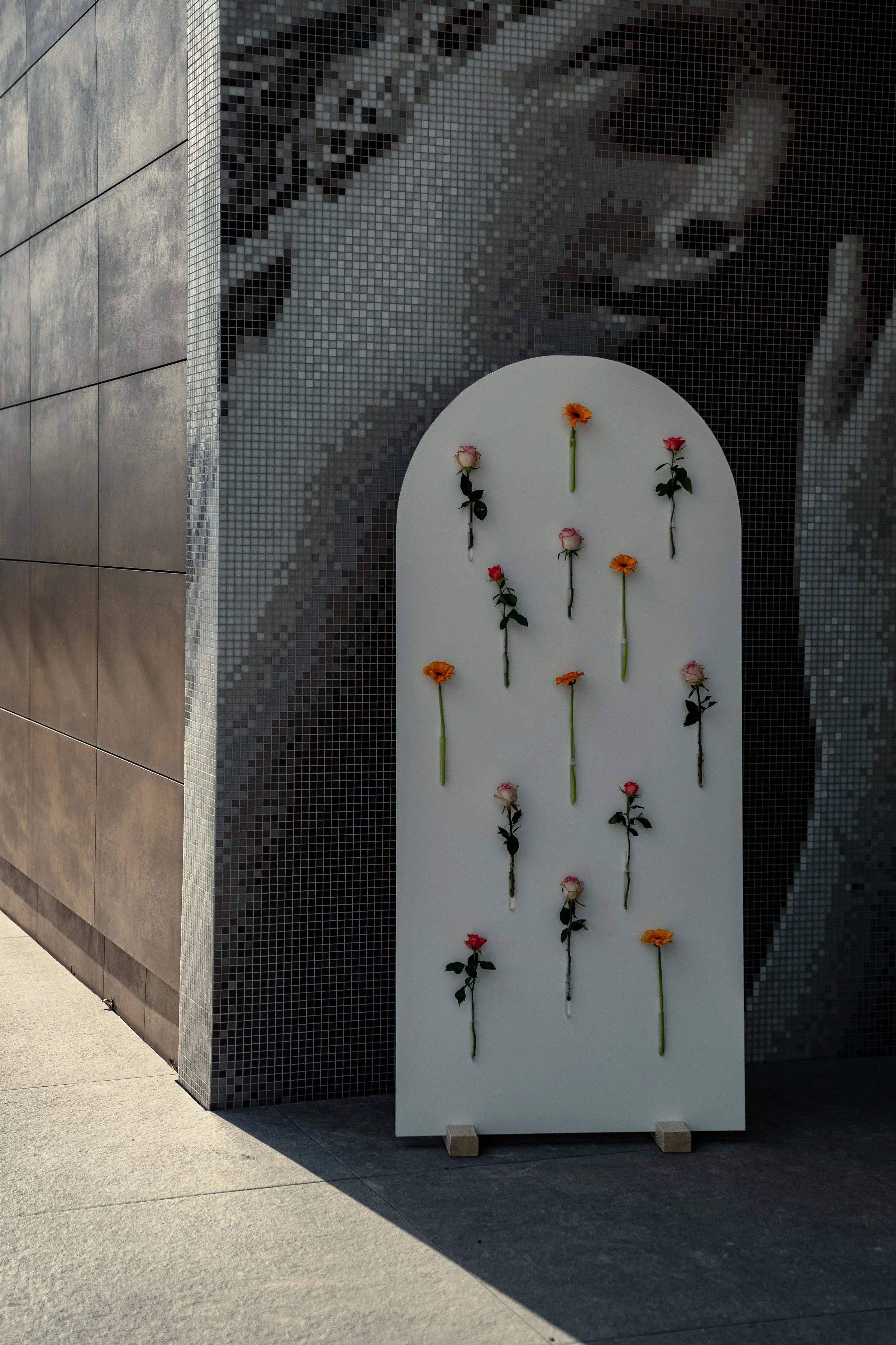 Ein weißer Rahmen mit aufgebrachten Rosen in verschiedenen Farben steht vor einer Mosaikwand, die ein Porträt zeigt.