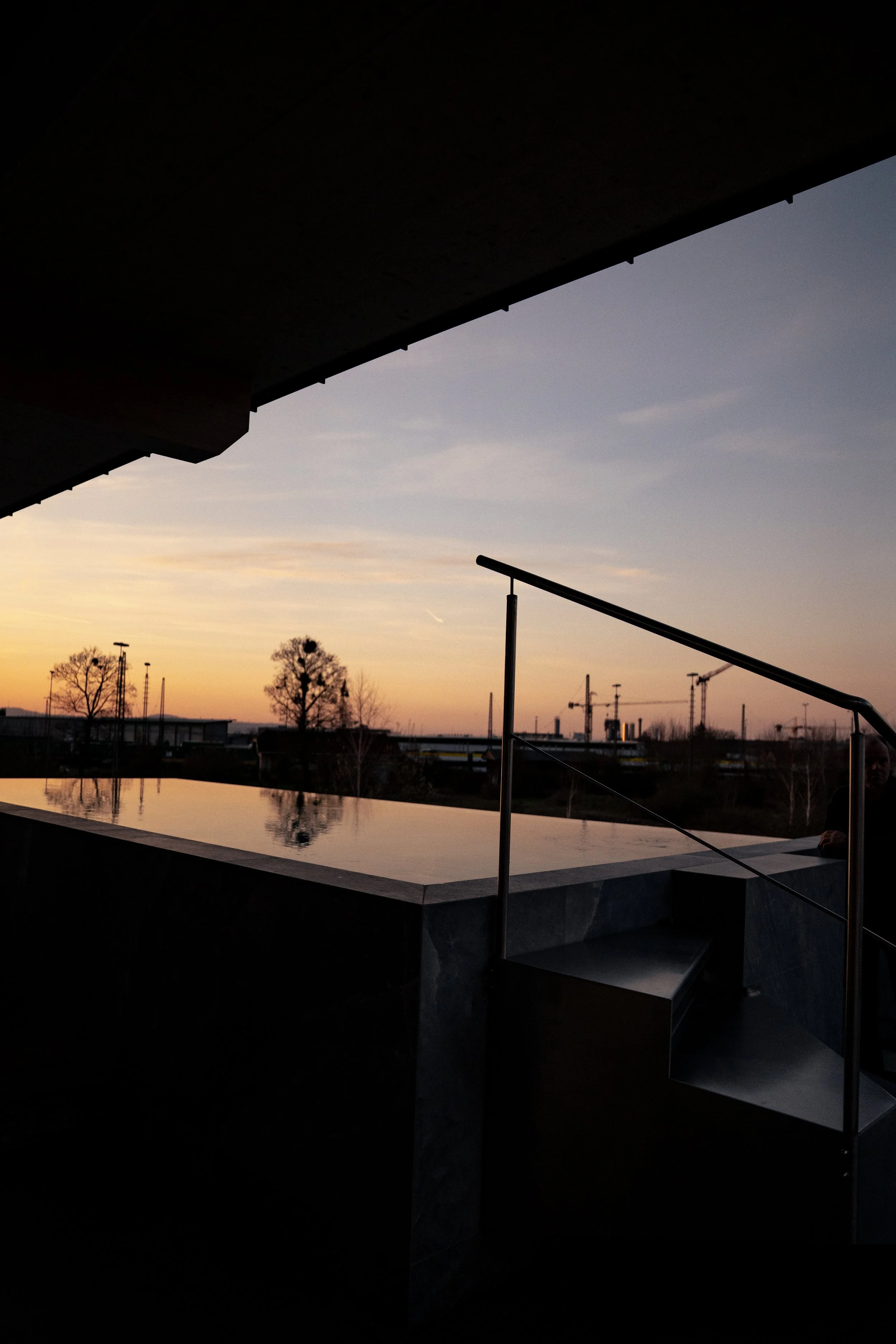 Blick auf einen Sonnenuntergang hinter einer modernen Gebäudefassade mit Wasserbecken und Stufen im Vordergrund.