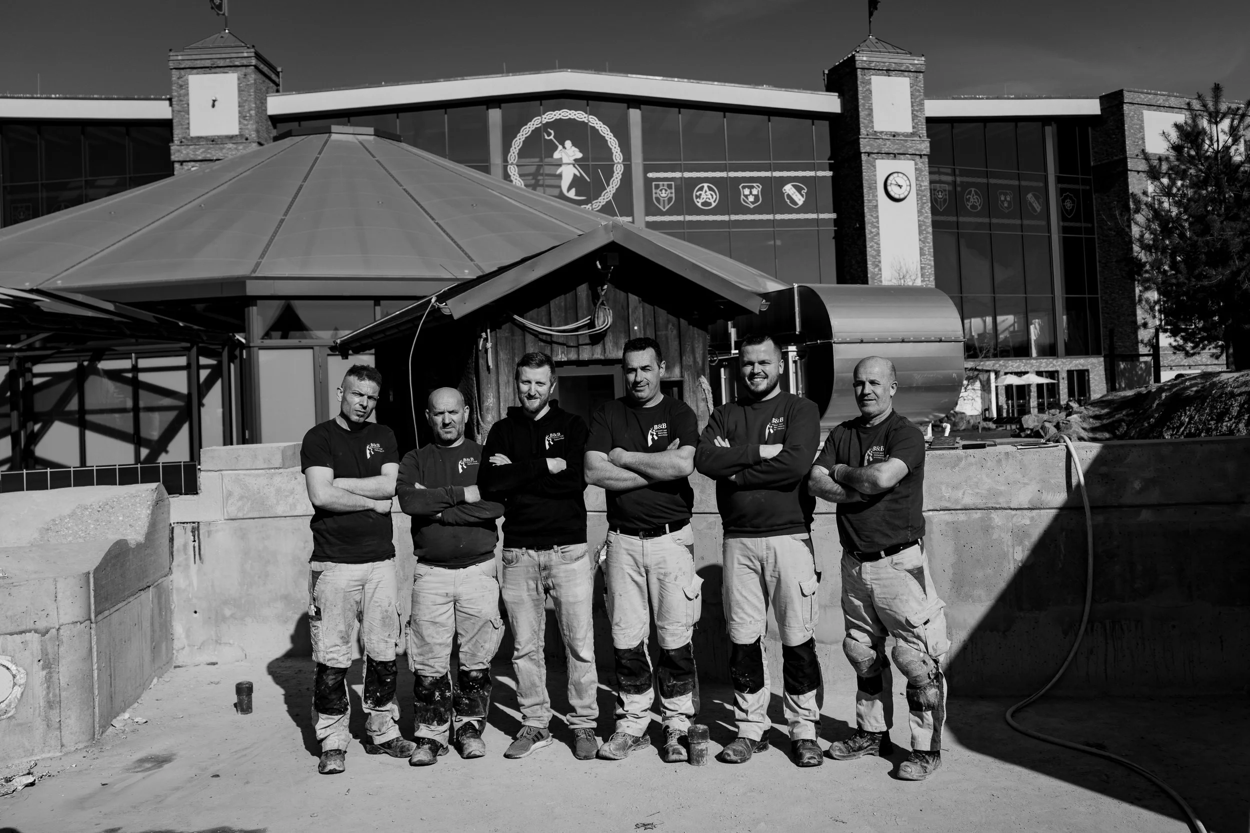 Sechs Bauarbeiter stehen mit verschränkten Armen vor einer Baustelle außerhalb eines modernen Gebäudes bei Sonnenlicht. Das Gebäude hat große Glasfenster, eine Uhr und auserlesene Symbole an der Fassade.