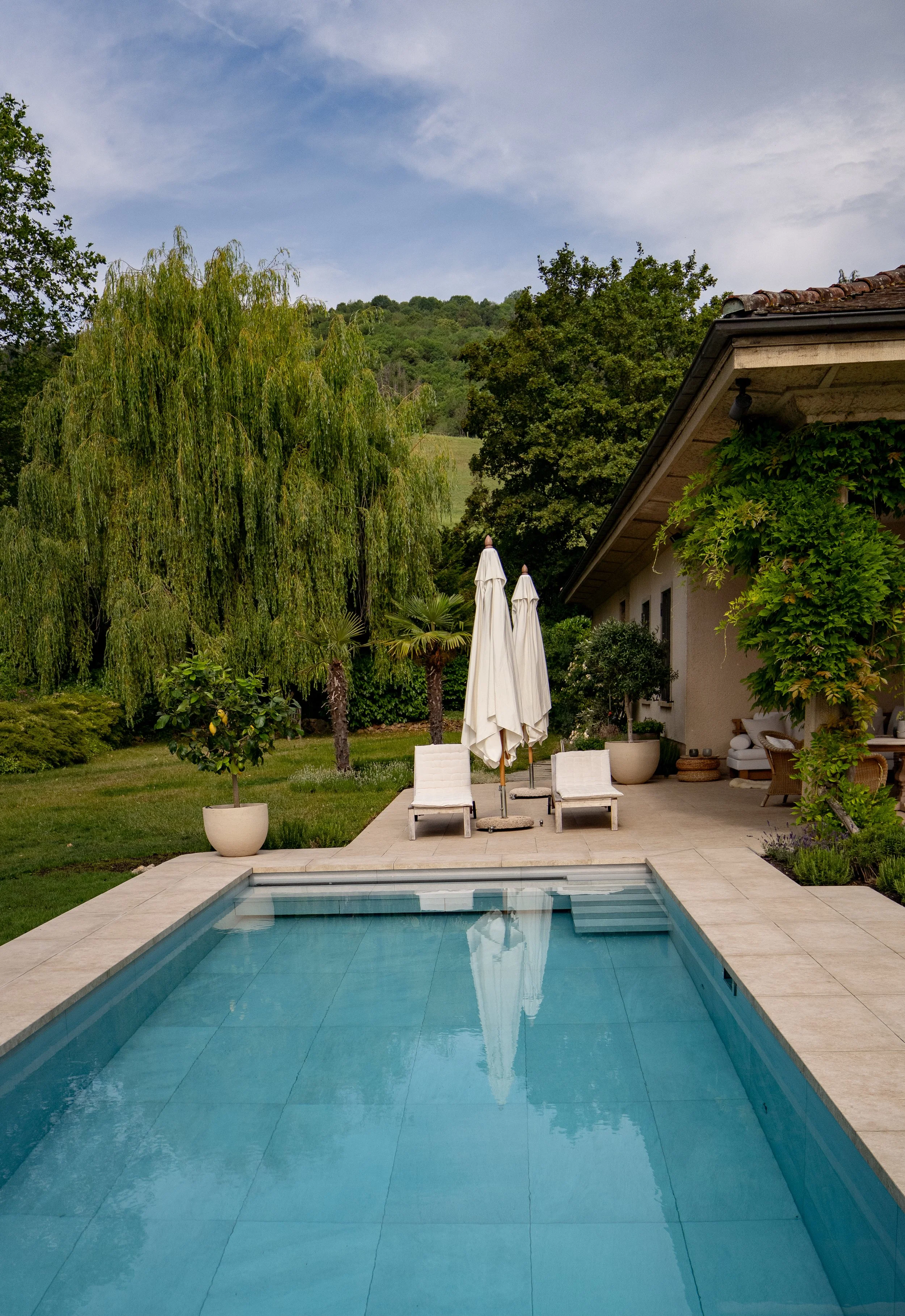 Ein Garten mit einem Swimmingpool, zwei Liegestühlen mit Sonnenschirmen, umgeben von Bäumen und einem Haus im Hintergrund.