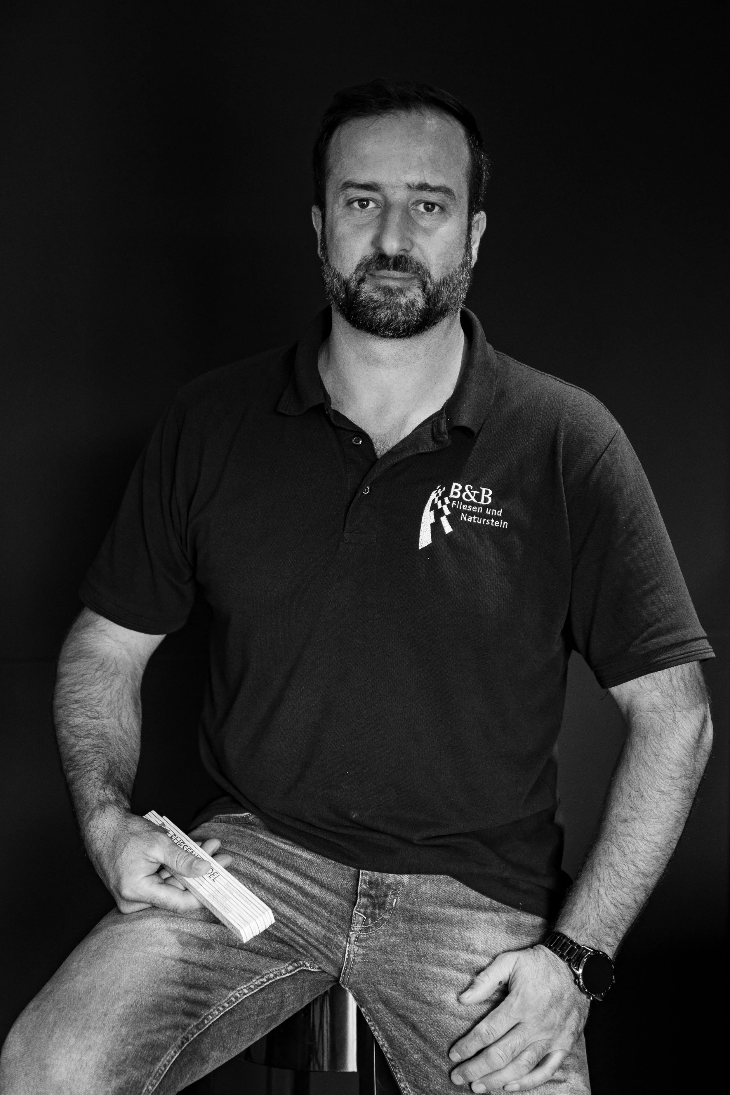 Ein Mann mit Bart sitzt auf einem Hocker vor einem schwarzen Hintergrund, trägt ein schwarzes Polo-Shirt mit Logo und Schriftzug, hält ein Paket Holzstöckchen in der Hand, trägt eine Armbanduhr.