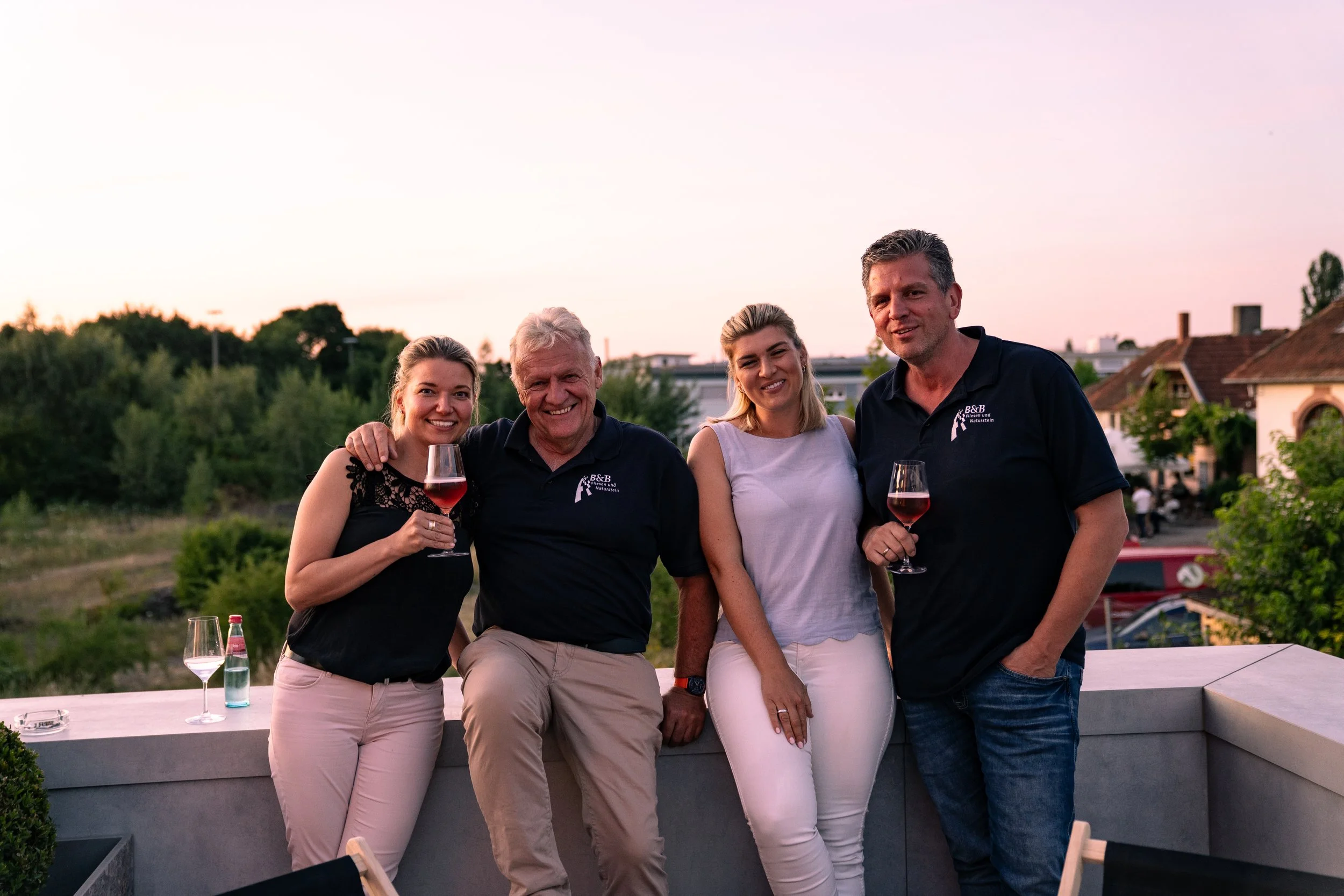 Vier Menschen auf einer Terrasse beim Zusammensitzen und Trinken von Rotwein bei Sonnenuntergang, mit Bäumen und Häuser im Hintergrund.