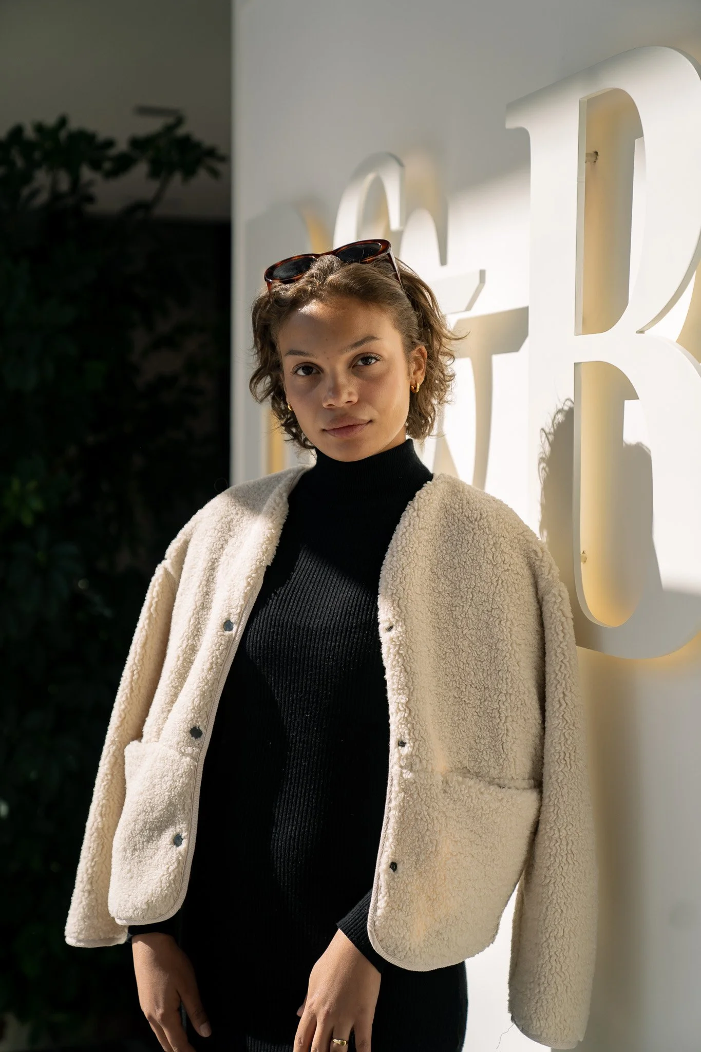 Eine junge Frau steht vor einer beleuchteten Wand mit großen weißen Buchstaben, trägt eine schwarze Rollkragenpullover und eine beige, kuschelige Jacke, Sonnenbrille auf dem Kopf, schaut direkt in die Kamera.