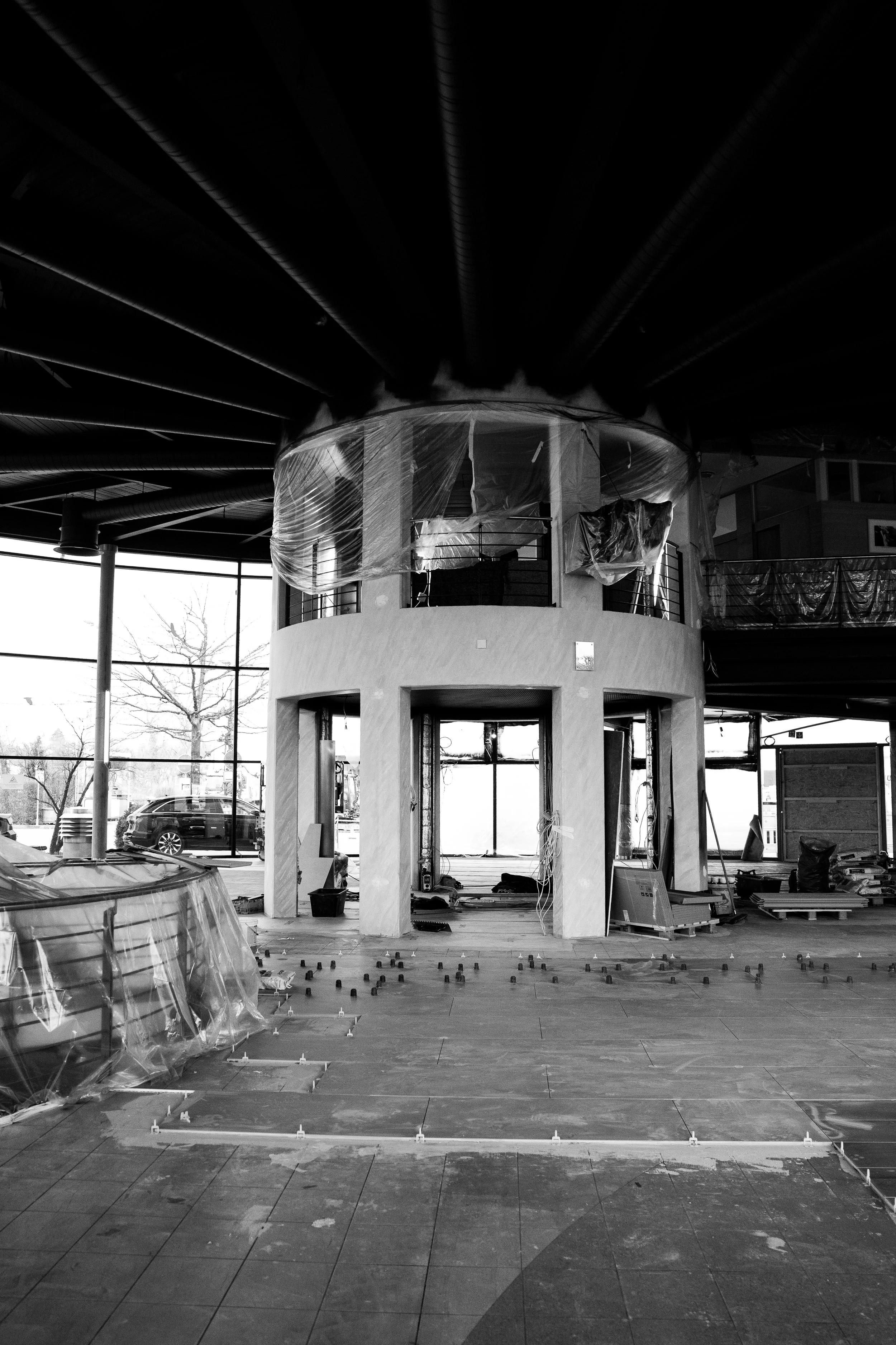 Innenraum eines Gebäudes unter Bau, mit sichtbar unvollendetem Obergeschoss, Baugeräte und Materialien, im Hintergrund große Fenster mit Blick nach draußen.