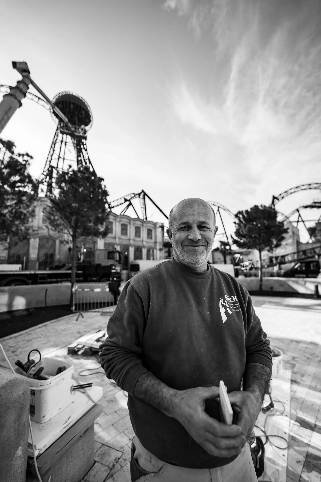 Ein älterer Mann steht vor einem Freizeitpark, trägt eine dunkle Sweatshirt mit Logo und hält einen Gegenstand in den Händen. Im Hintergrund sind Fahrgeschäfte, Bäume und Bauarbeiten zu sehen.