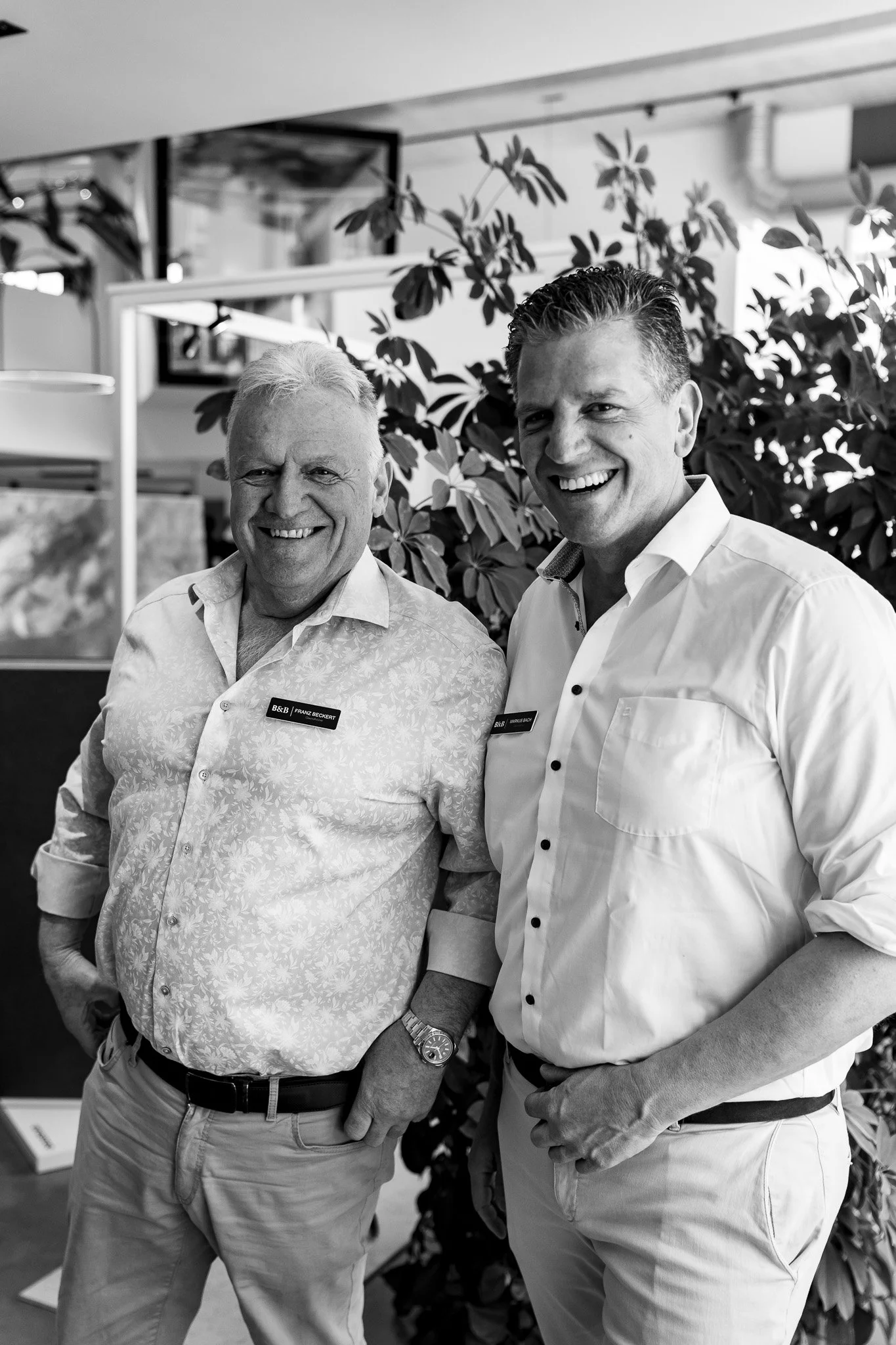 Zwei lächelnde Männer in Hemden, im Büro mit Pflanzen im Hintergrund.