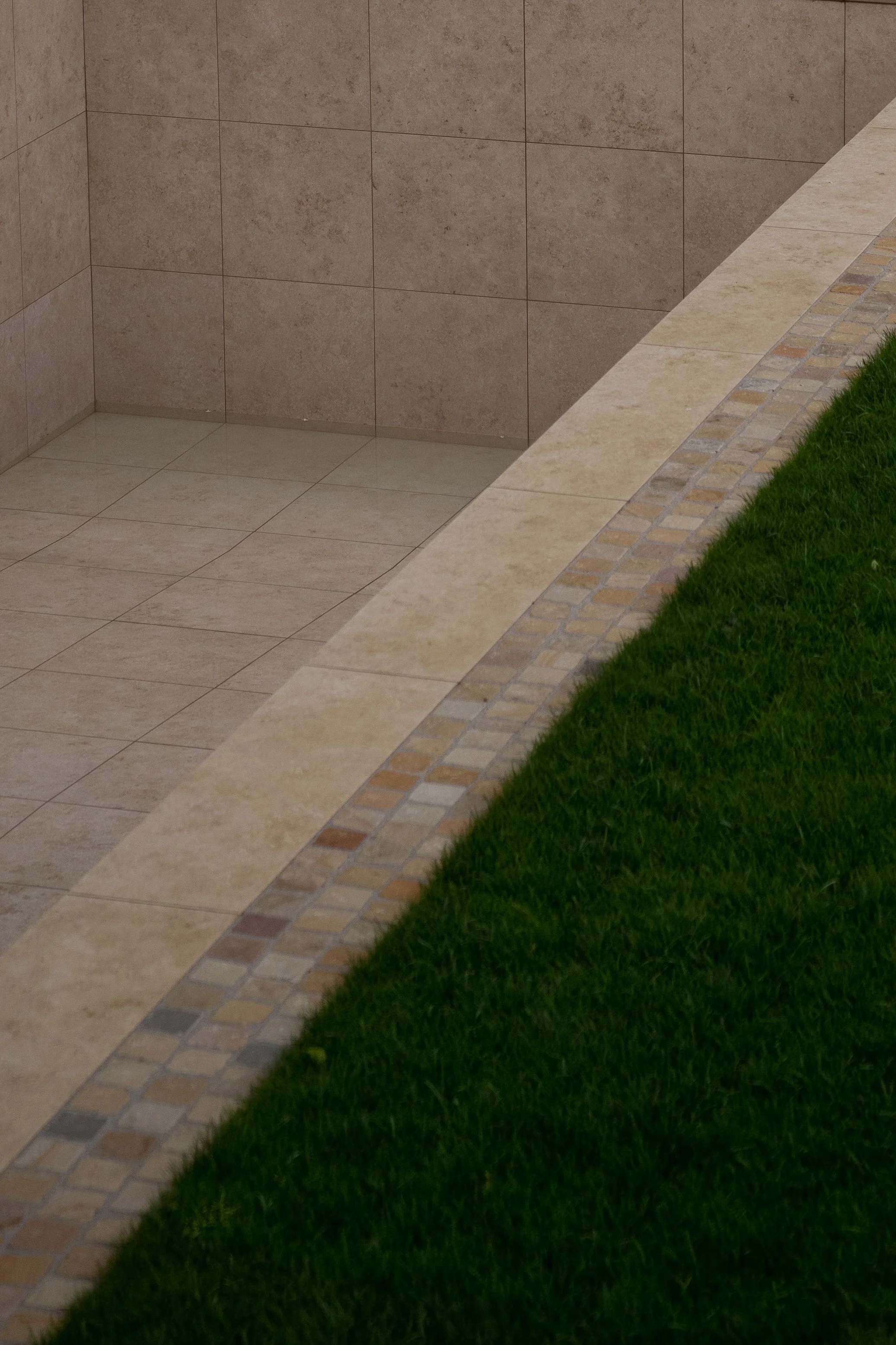 Blick auf die Ecke eines Außenbereichs mit Bodenfliesen, einer Bordüre mit kleinen Steinen und einem Rasen.