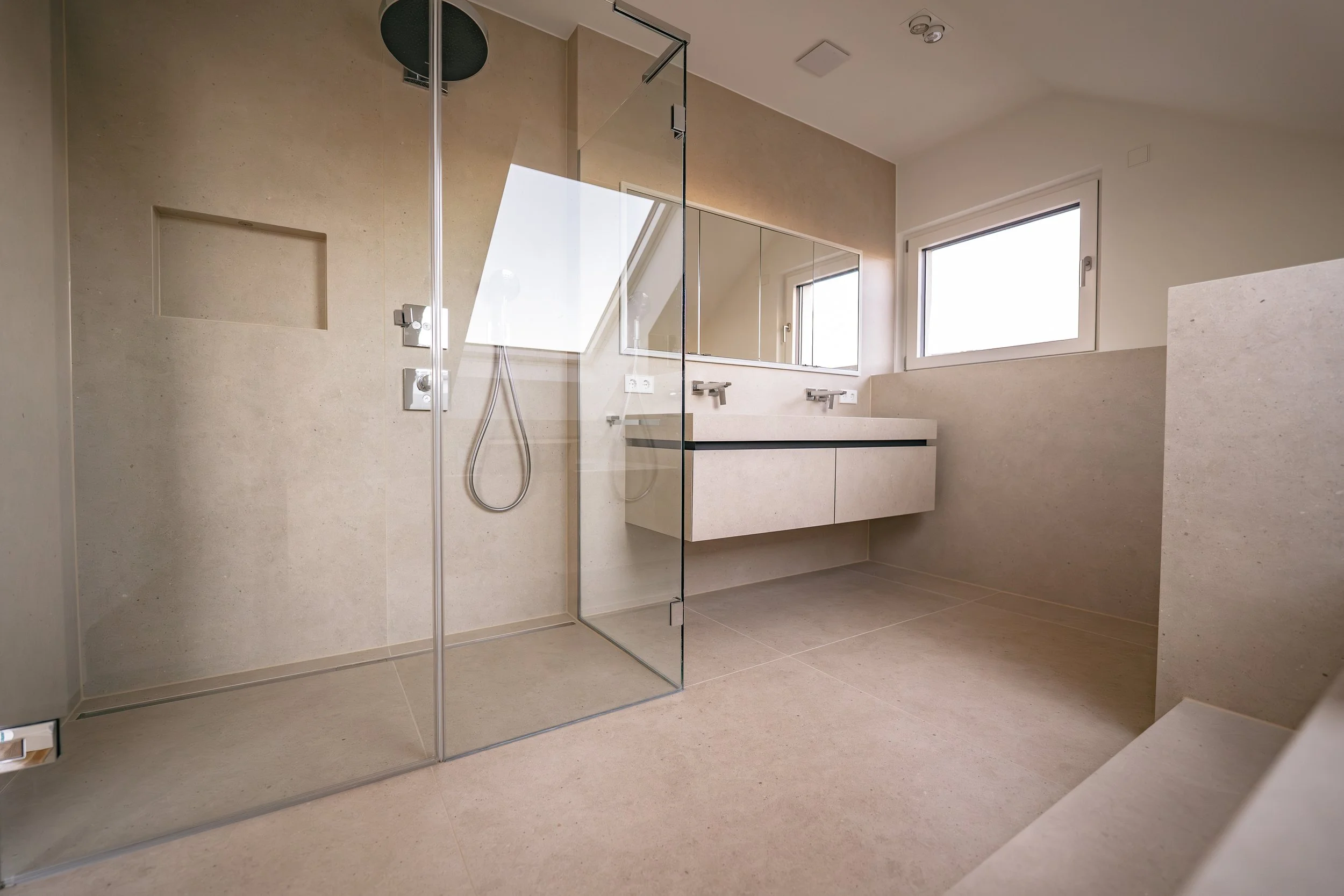 Modernes Badezimmer mit beiger Stein-Design, eine ebenerdige Dusche mit Glasabtrennung, Doppelwaschbecken, große Fenster und minimalistische Einrichtung.