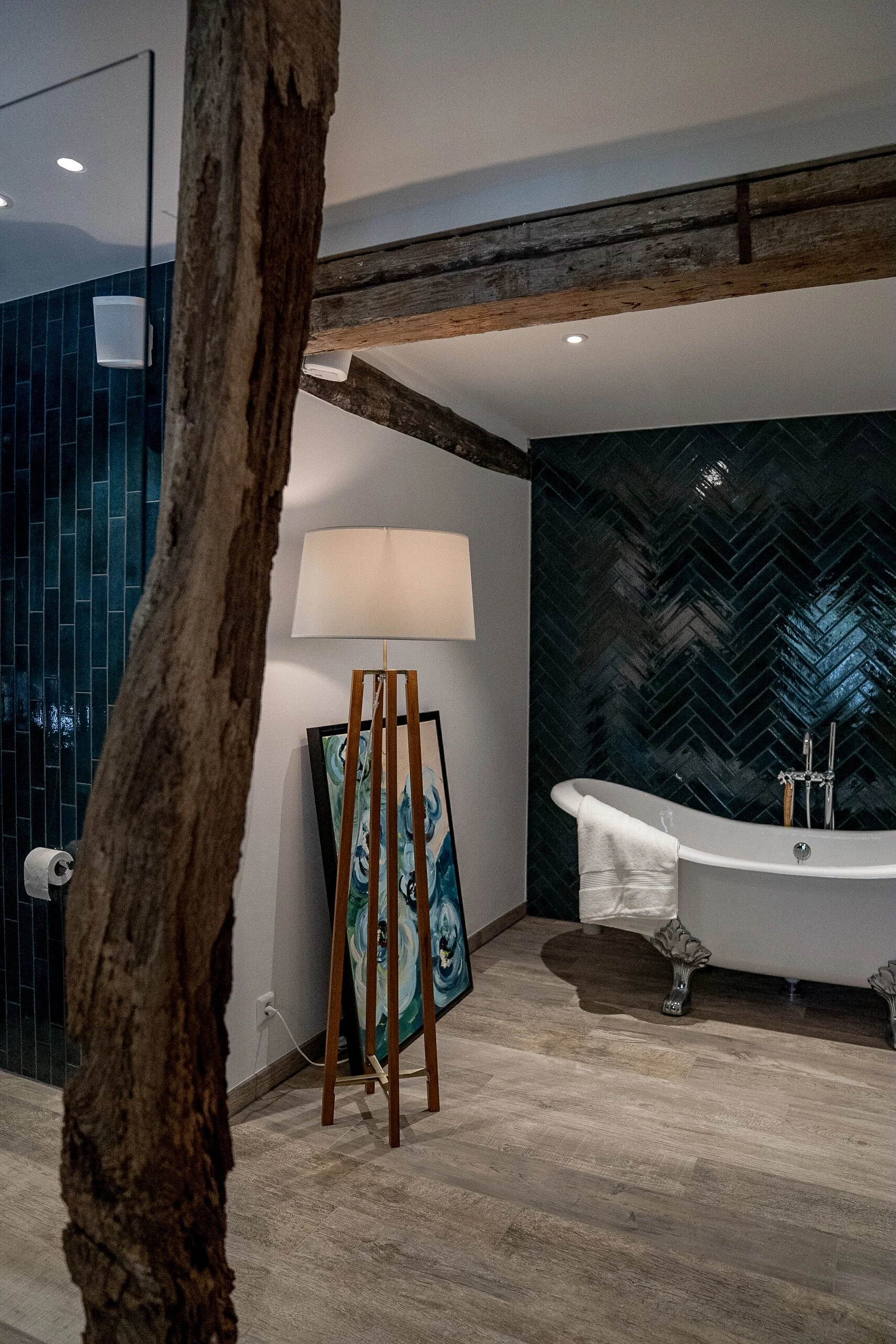 Elegant Badezimmer mit freistehender Badewanne, dunklen Fliesen hinter der Badewanne, Holzdecke mit sichtbaren Balken, Stehlampe und moderner Kunst im Raum.