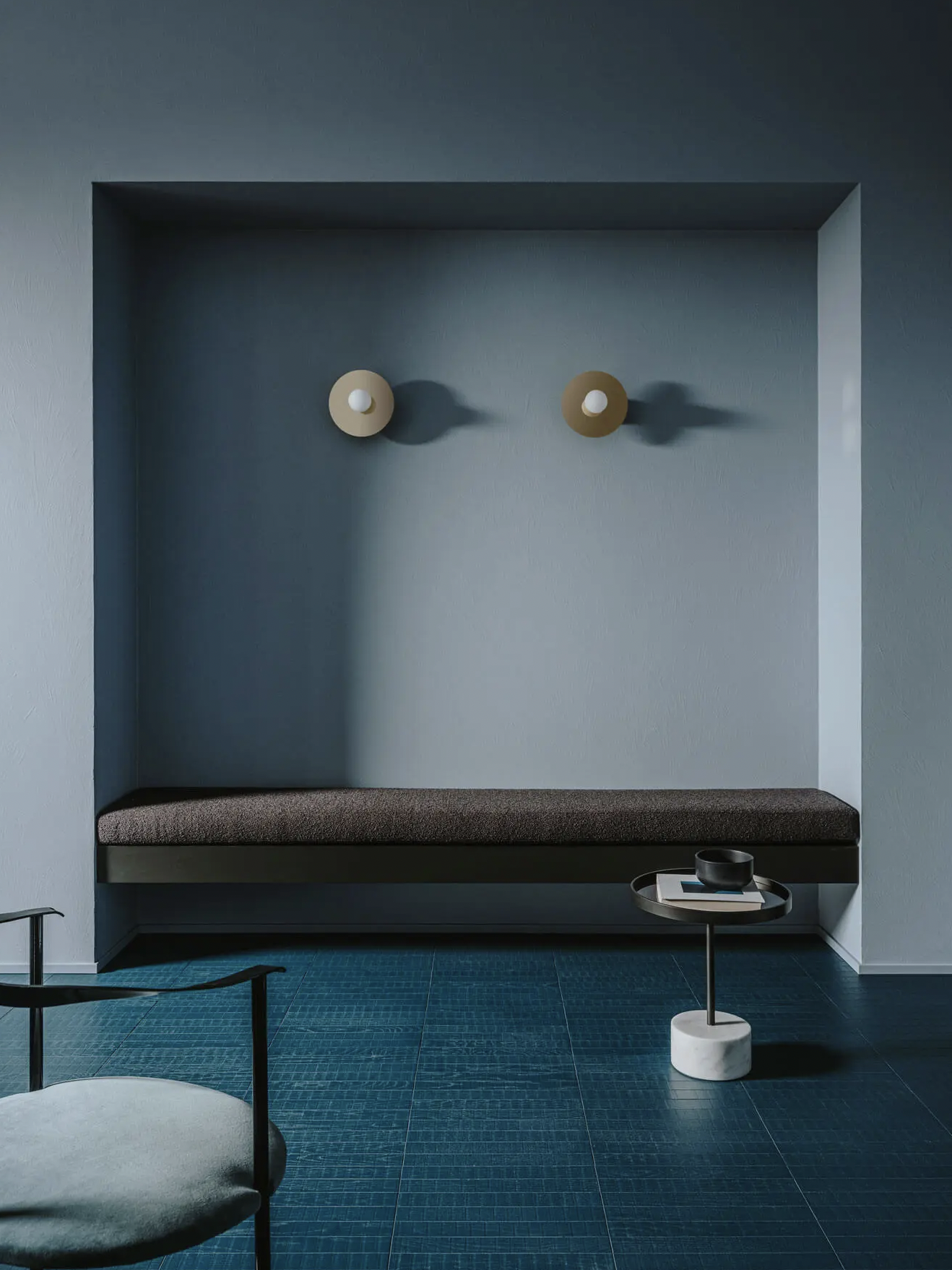 Ein moderner, minimalistisch gestalteter Eingangsbereich mit dunkelblauer Wand und Sitzbank. Zwei runde Wandlampen werfen Schatten, und ein kleiner runder Beistelltisch mit Gegenständen steht auf dem Boden.