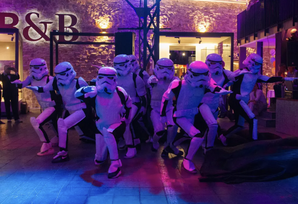 Menschen in Stormtrooper-Kostümen tanzen auf einer Party mit bunter Beleuchtung in einem Raum mit Steinen an der Wand.