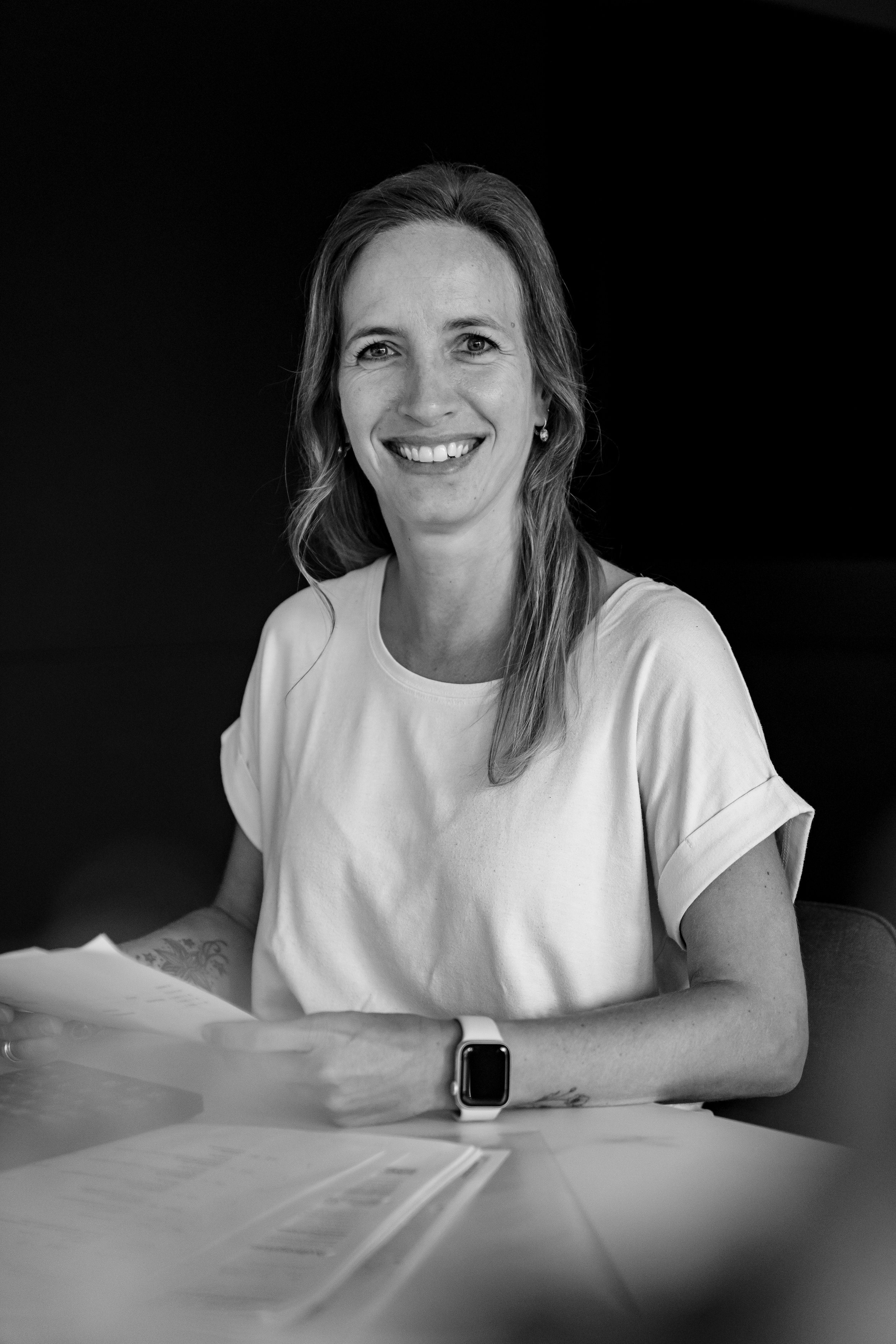 Schwarz-weiße Fotografie einer lächelnden Frau mit langen Haaren, die in einem weißen T-Shirt sitzt, neben einem Handy und Papieren.