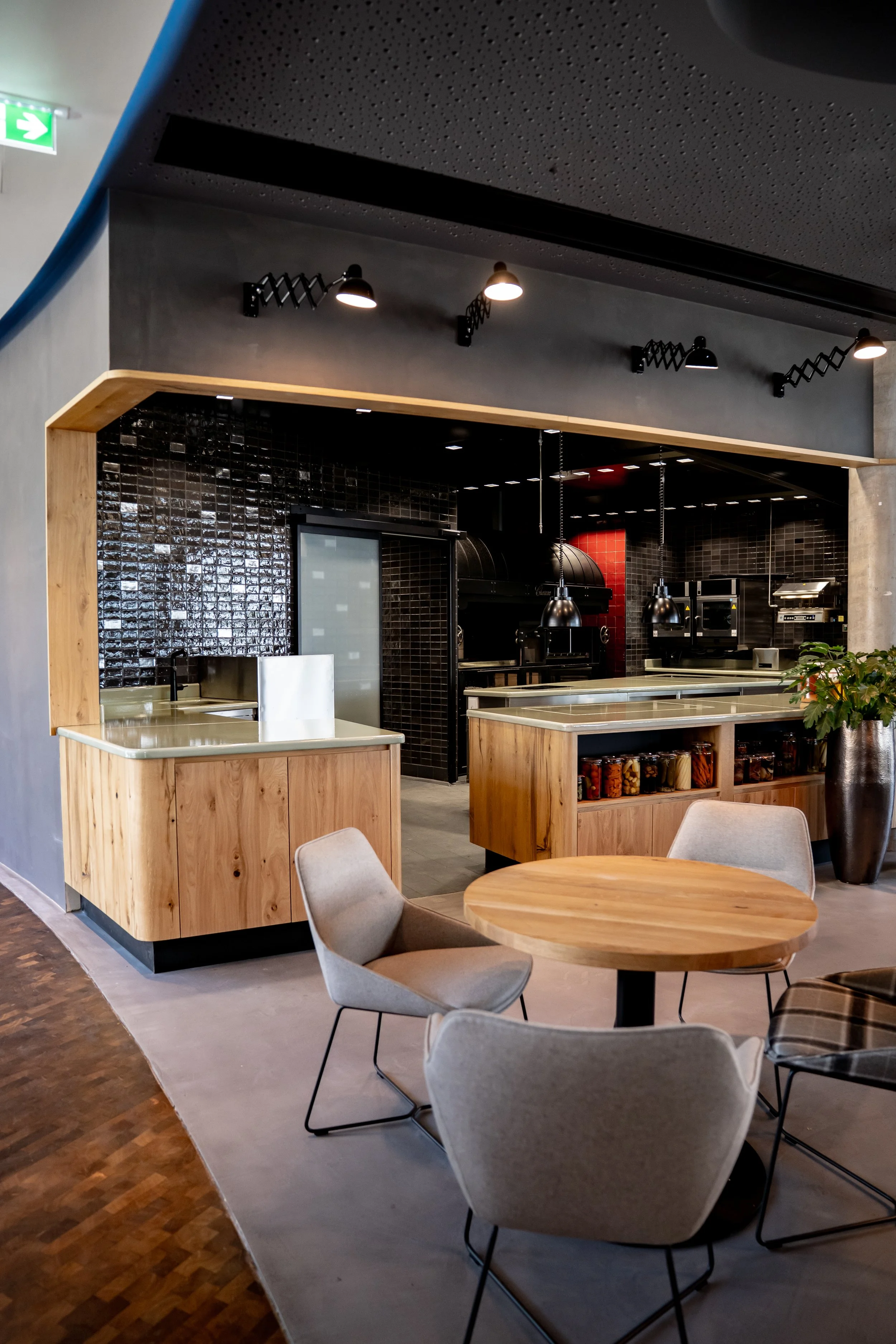 Moderne Restaurantküche mit dunklen Fliesen, Holzarbeitsflächen, Küchengeräten und gemütlichem Sitzbereich mit Stühlen und Tisch.