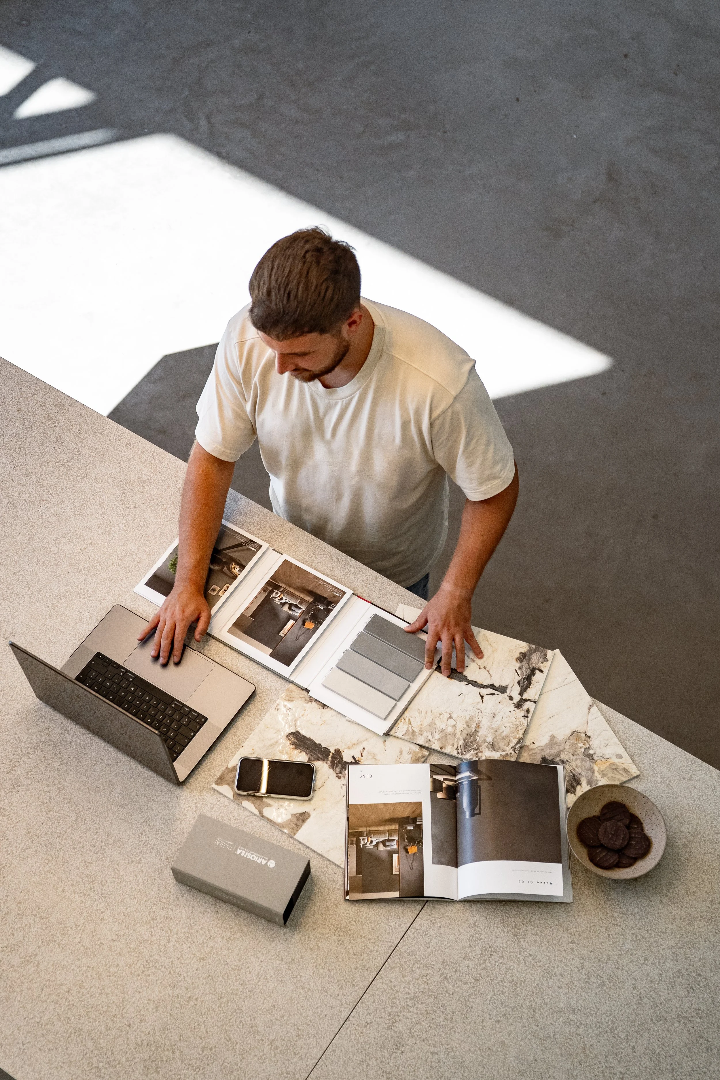 Ein Mann steht an einem Tisch mit einem Laptop, Design- und Farbmustern, gedruckten Materialproben, einem Telefon, einem Paket und einer Schüssel Kekse, während er durch Design- oder Einrichtungspläne blättert.
