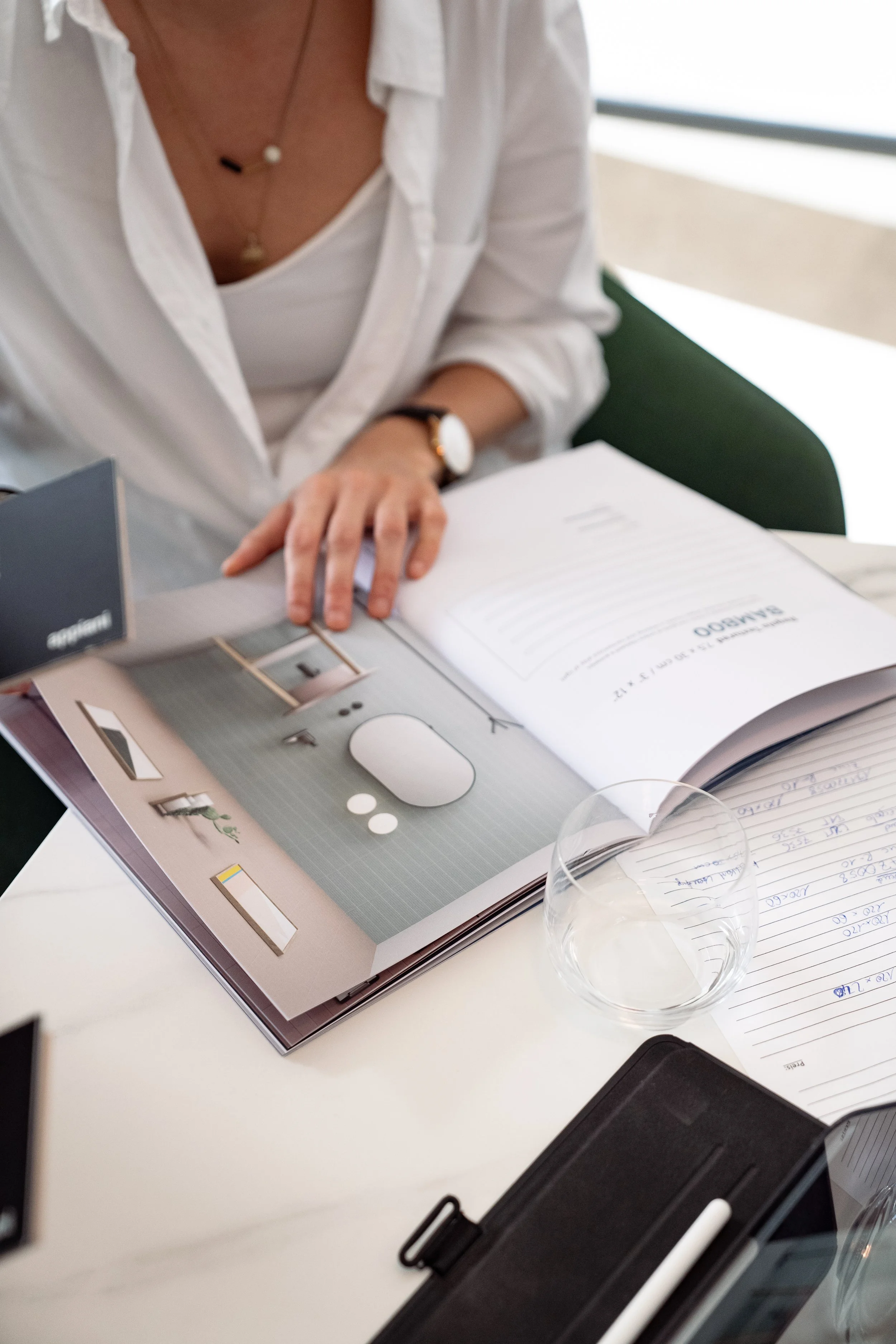 Eine Person liest ein aufgeschlagenes Buch oder Magazin an einem Schreibtisch, umgeben von Notizen, einem leeren Glas, einer schwarzen Mappe mit Stift und anderen Büromaterialien.