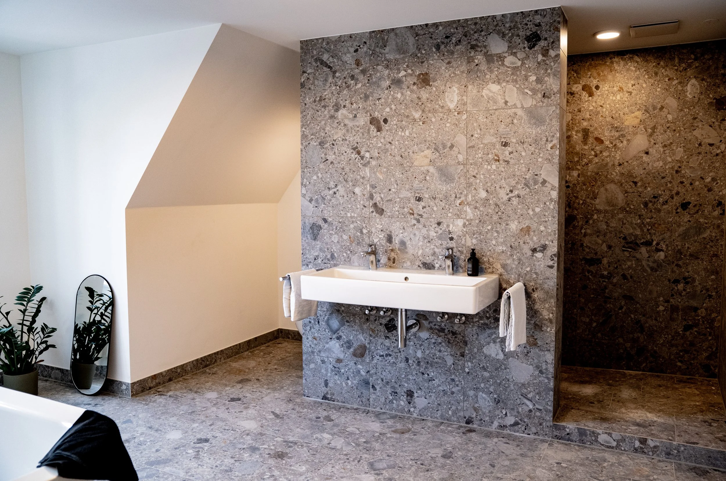 Badezimmer mit Wandwaschbecken und grauer Marmorwand, dunkelbraune Duschwand im Hintergrund, Pflanzen und Spiegel im linken Bereich.