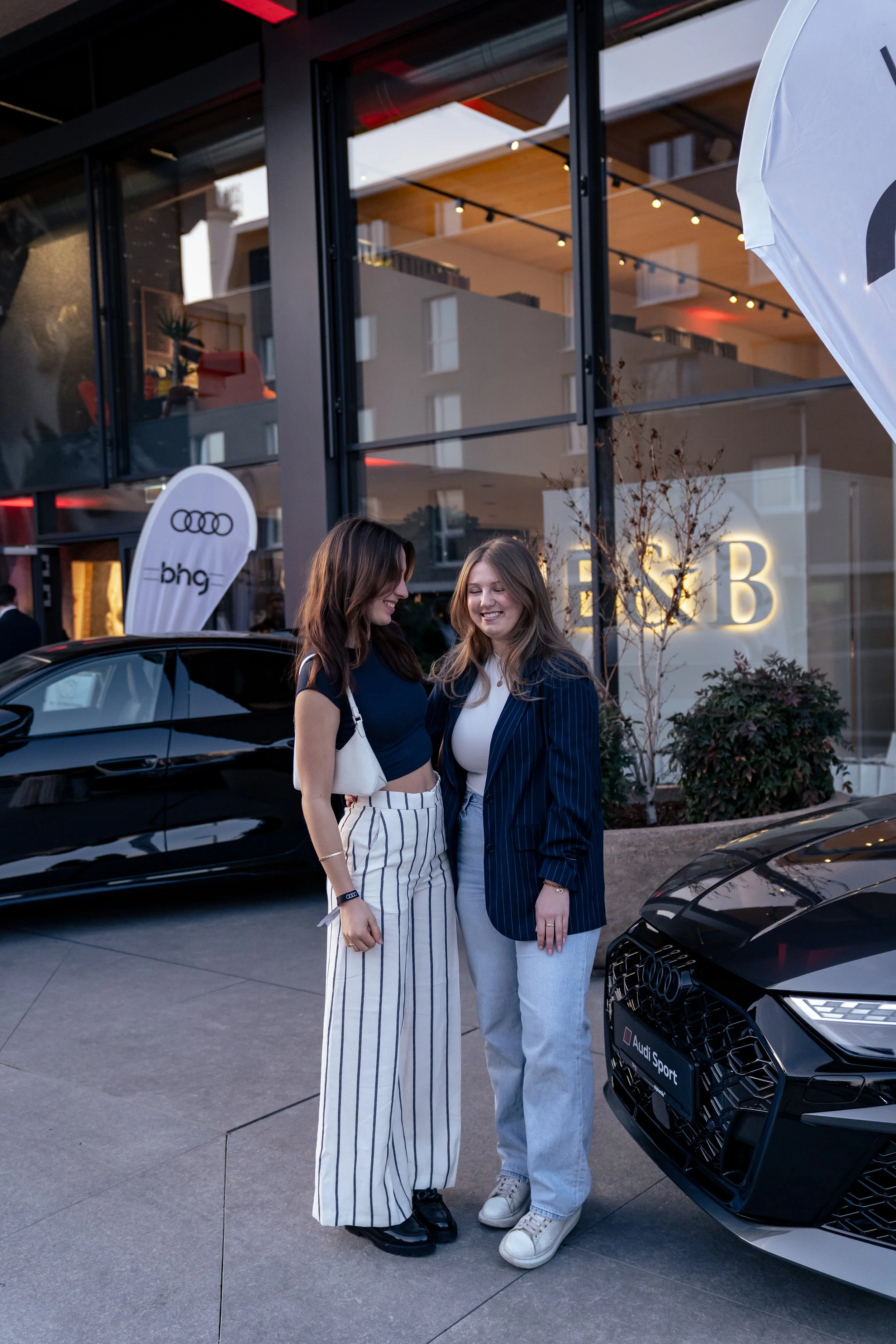 Zwei Frauen stehen lächelnd vor einem schwarzen Audi Sport Auto bei einer Veranstaltung außerhalb eines eleganten Gebäudes.