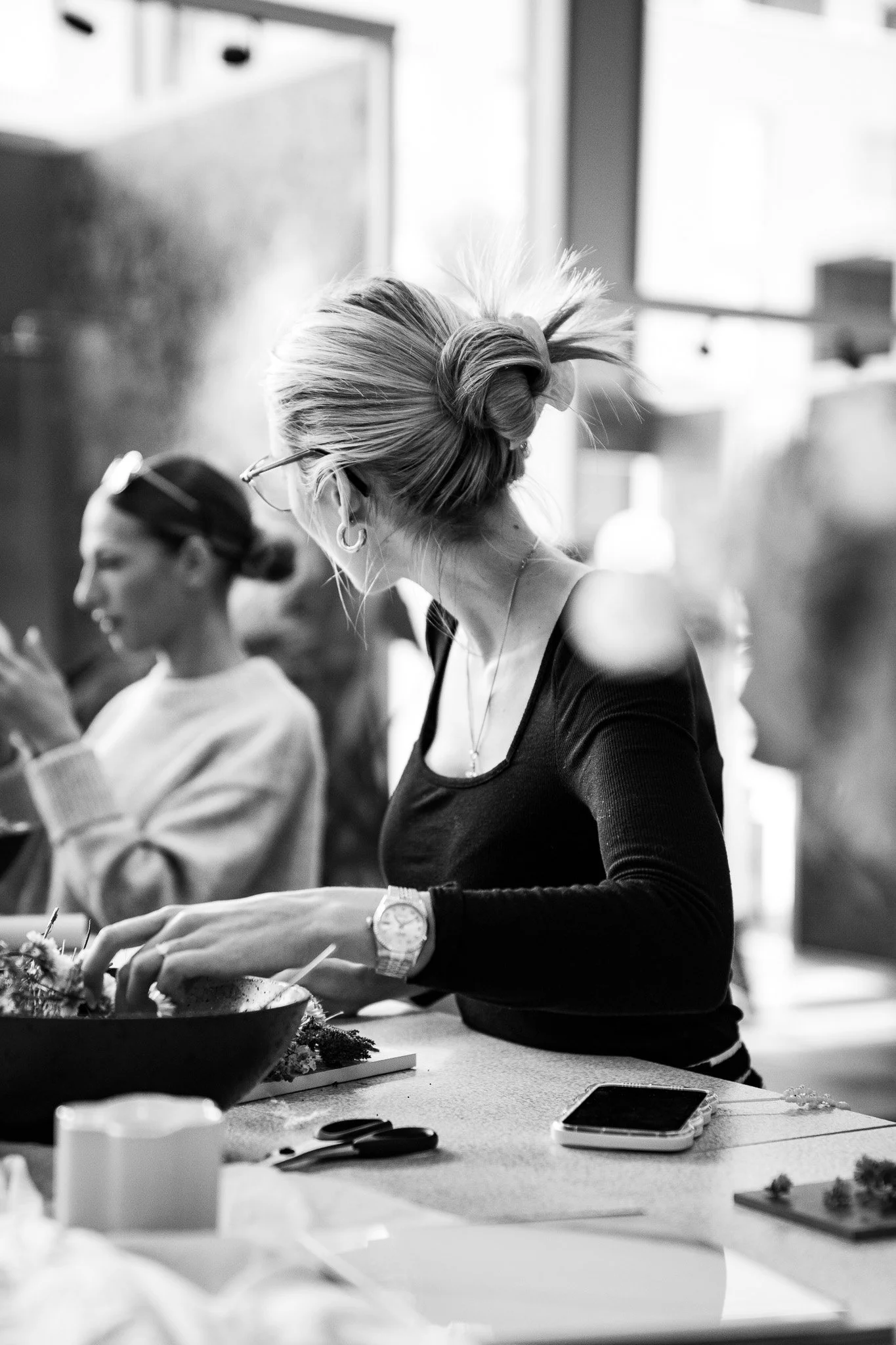 Schwarze und weiße Aufnahme einer Gruppe von Frauen, die an einem Tisch sitzen, wobei die Frau im Vordergrund eine Armbanduhr trägt und beim Essen ist.