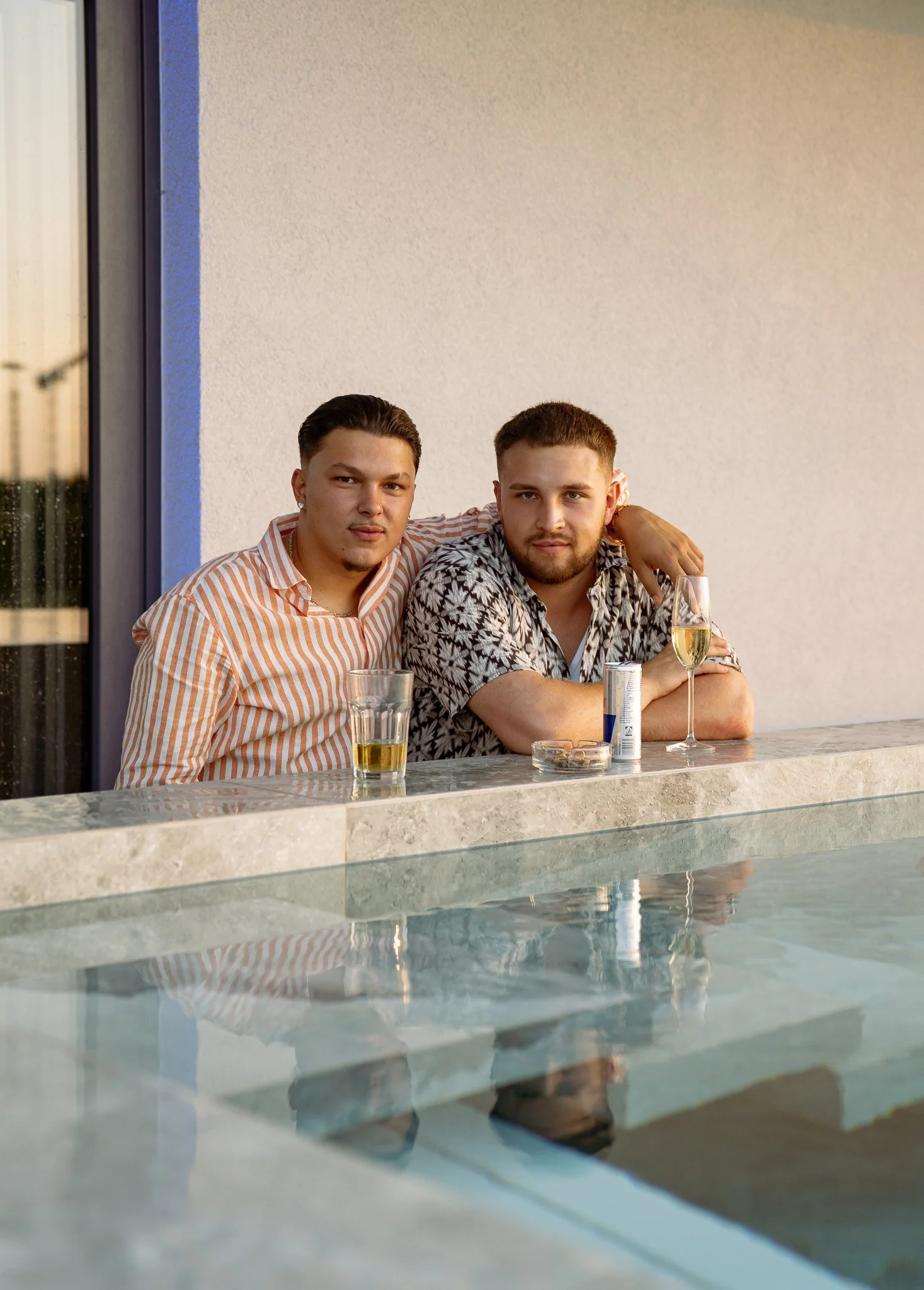 Zwei Männer sitzen an einem Außenbereich mit Blick auf einen Pool, trinken Getränke und schauen in die Kamera.