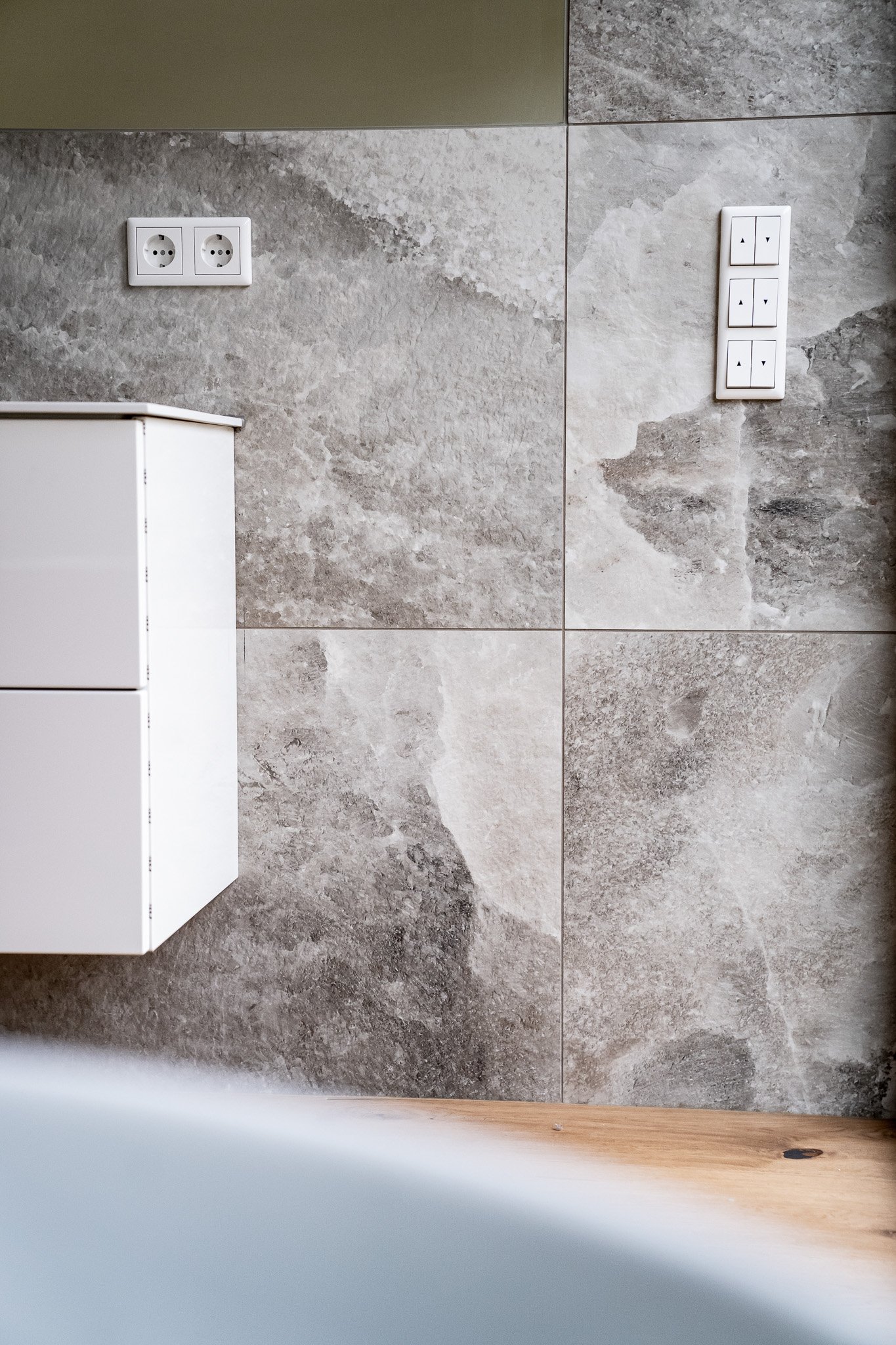 Teil einer Wand mit grauen Fliesen, zwei europäische Steckdosen, eine vertikale Reihe von sechs Lichtschaltern, eine weiße kleine Schublade und ein Holzboden