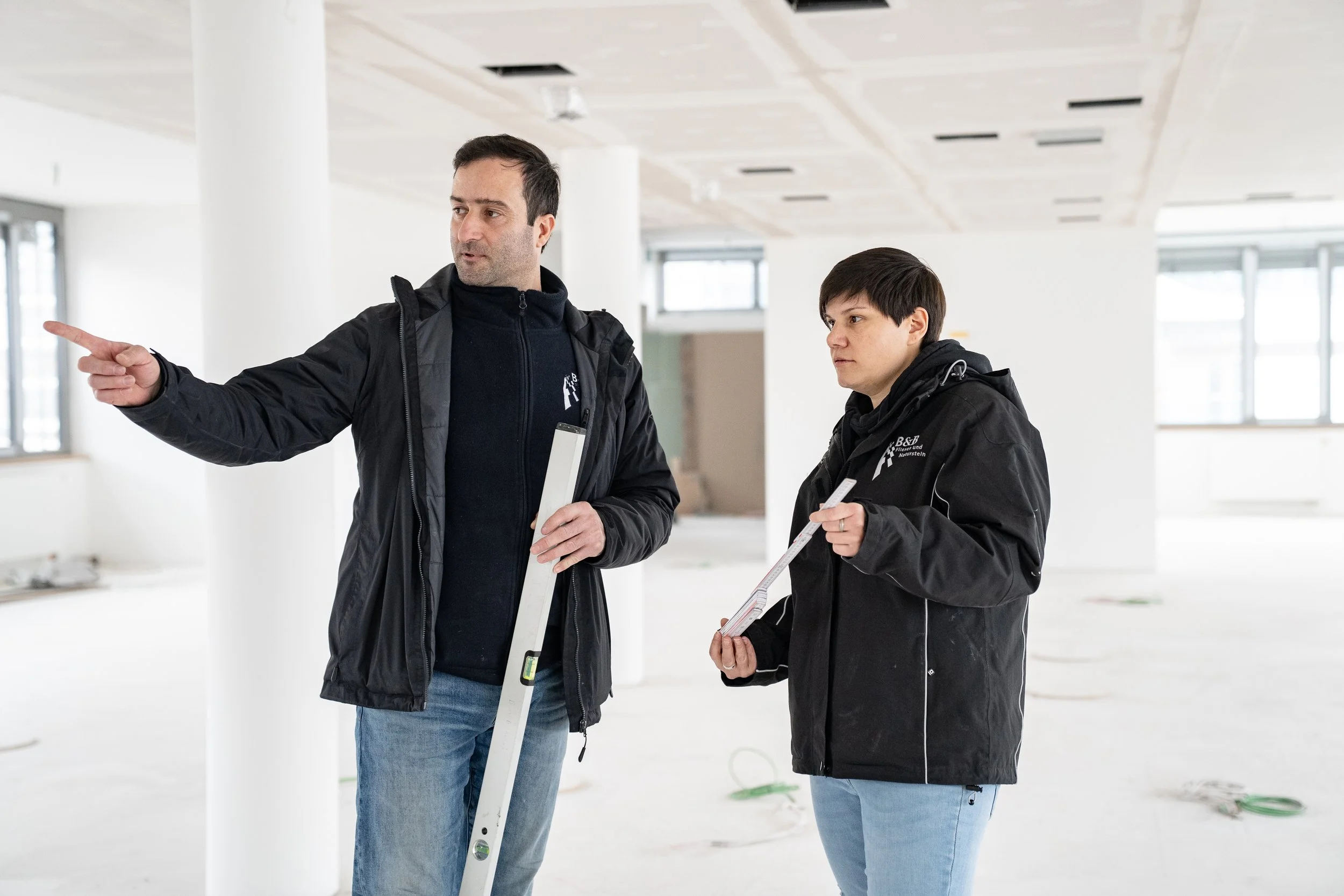 Zwei Bauleiter besprechen sich in einem unfertigen Gebäude, eines zeigt mit dem Finger auf einen Punkt, während das andere Notizen hält.