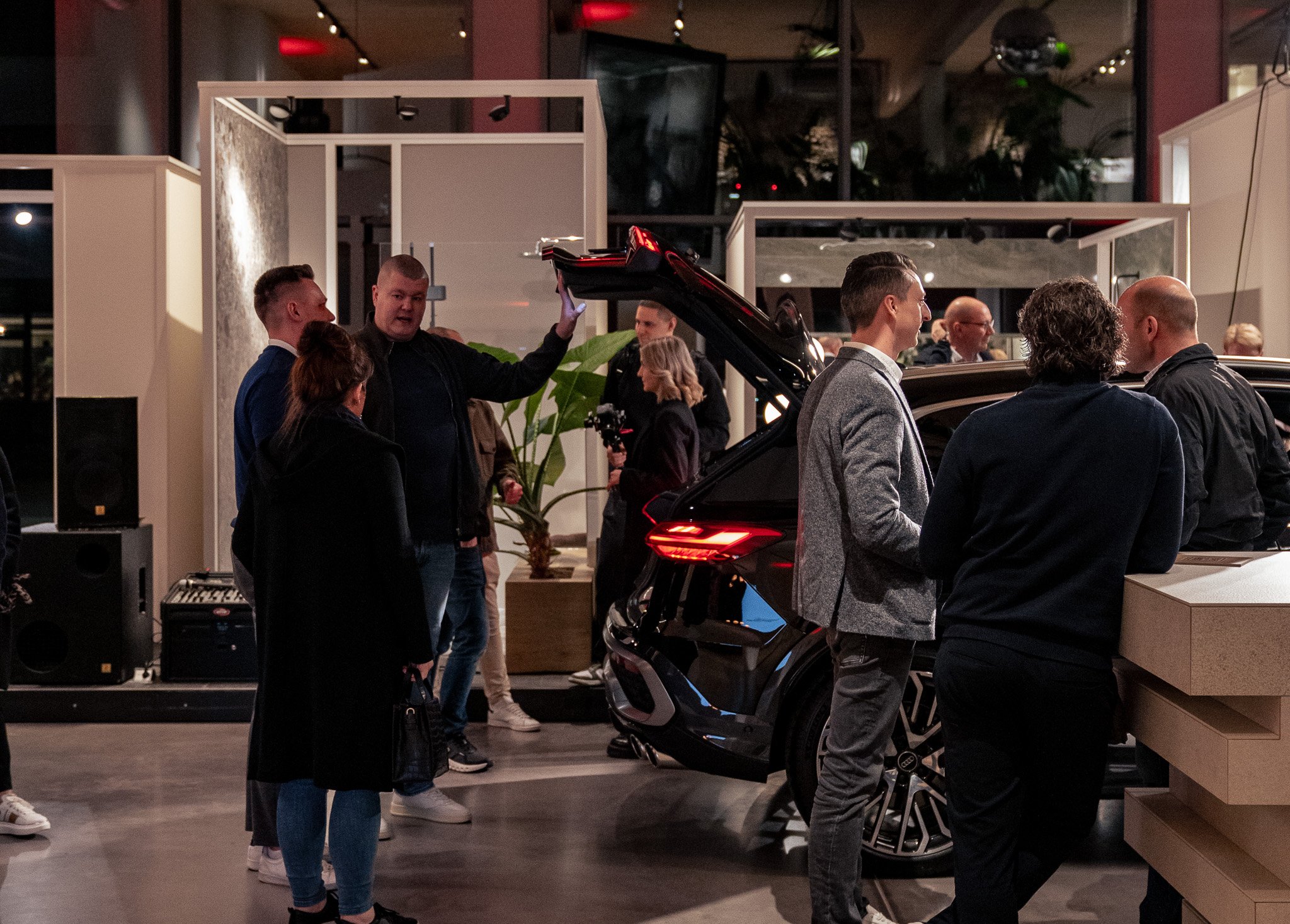 Menschen beim Autokauf oder -präsentation in einem Raum mit einem schwarzen Fahrzeug, das geöffnet ist, und einer Person, die das Fahrzeug erklärt.