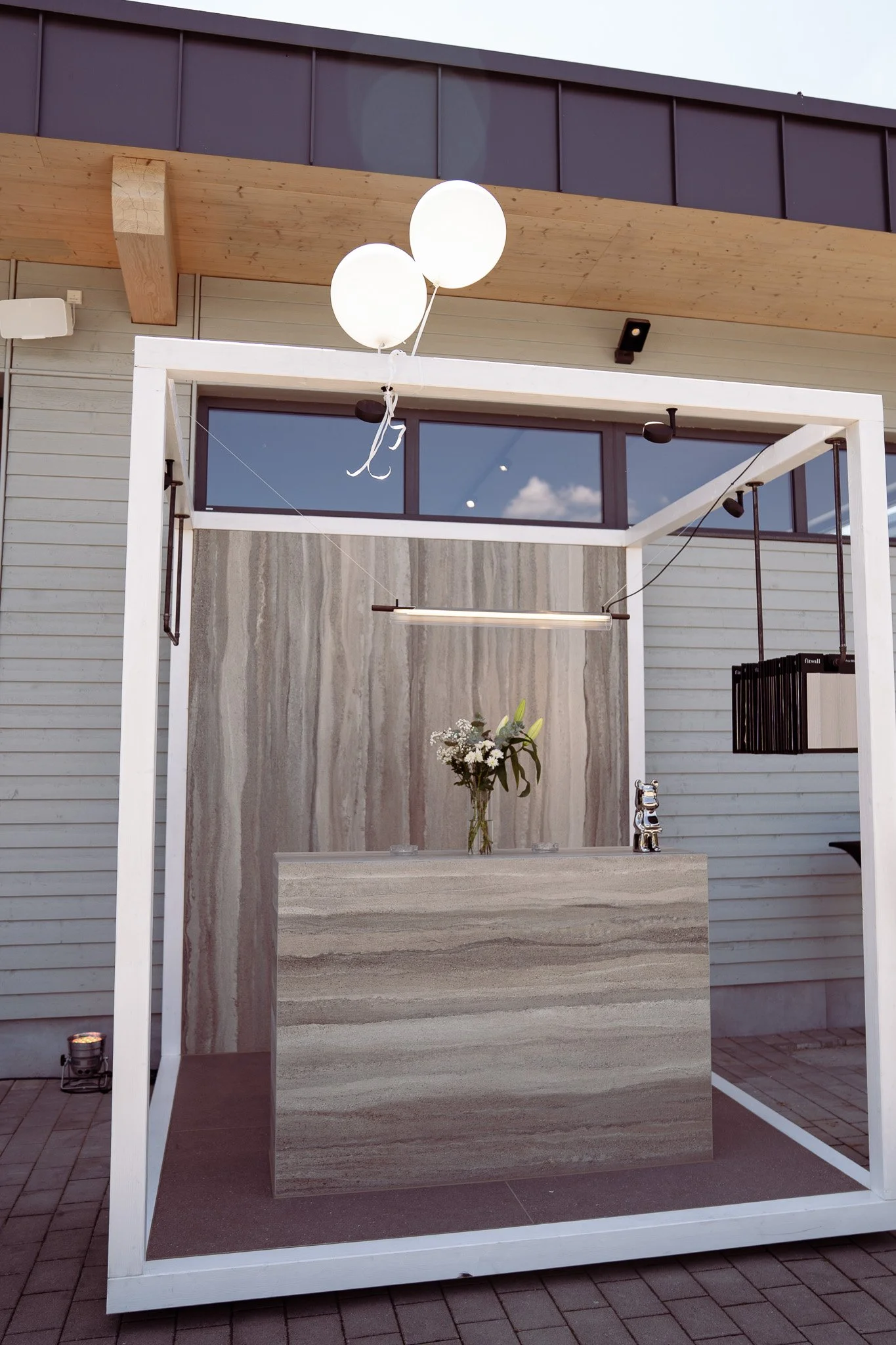 Ein moderner, offener Pavillon mit einer Theke aus marmorartigem Stein, einer Vase mit weißen Blumen und schwarzen Ballons, im Freien vor einem Gebäude mit Holz- und Glasfassade.