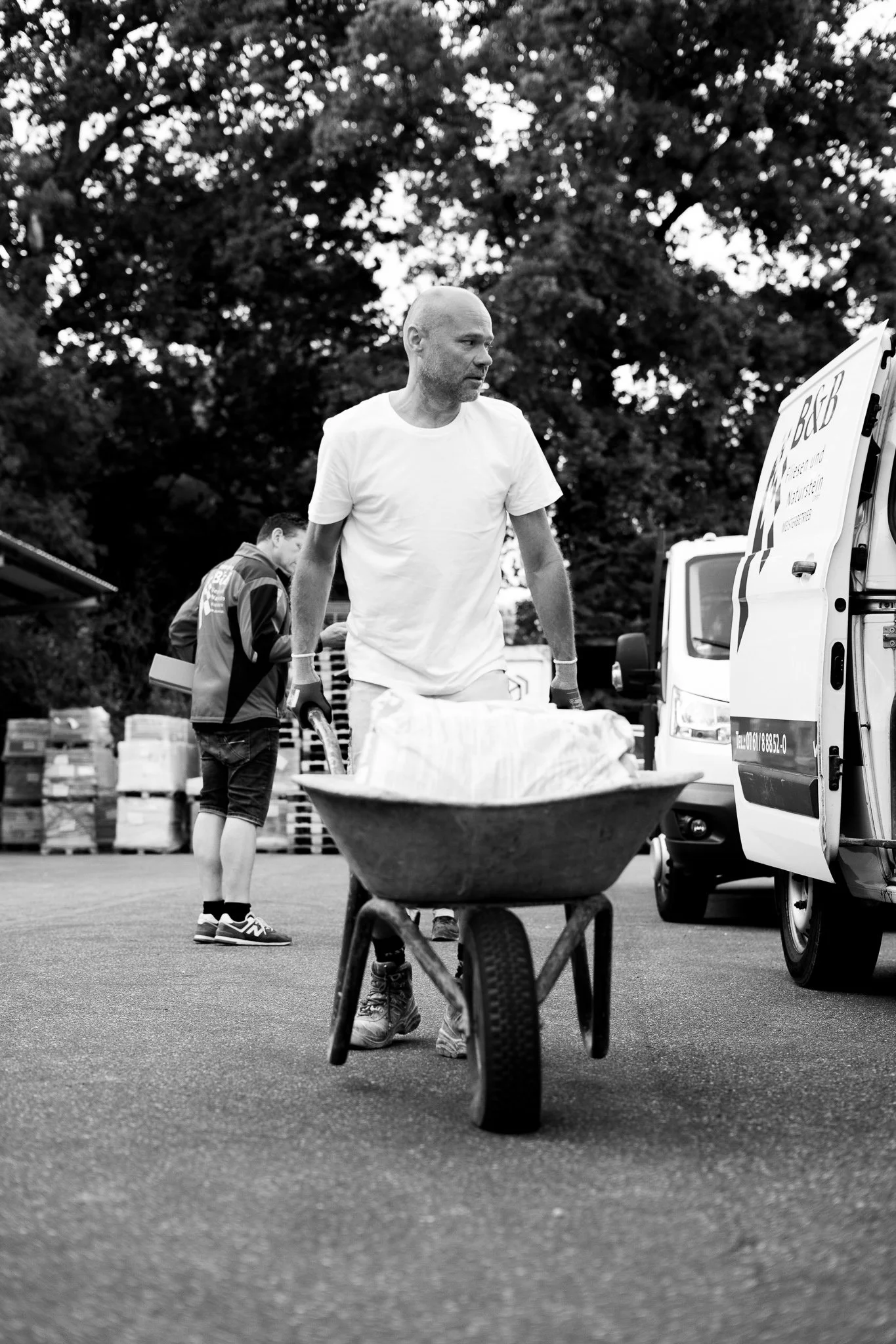 Mann schiebt einen Handkarren mit einem großen Gegenstand, im Hintergrund weitere Personen und Fahrzeuge, sie scheinen bei einer Arbeit im Freien zu sein.