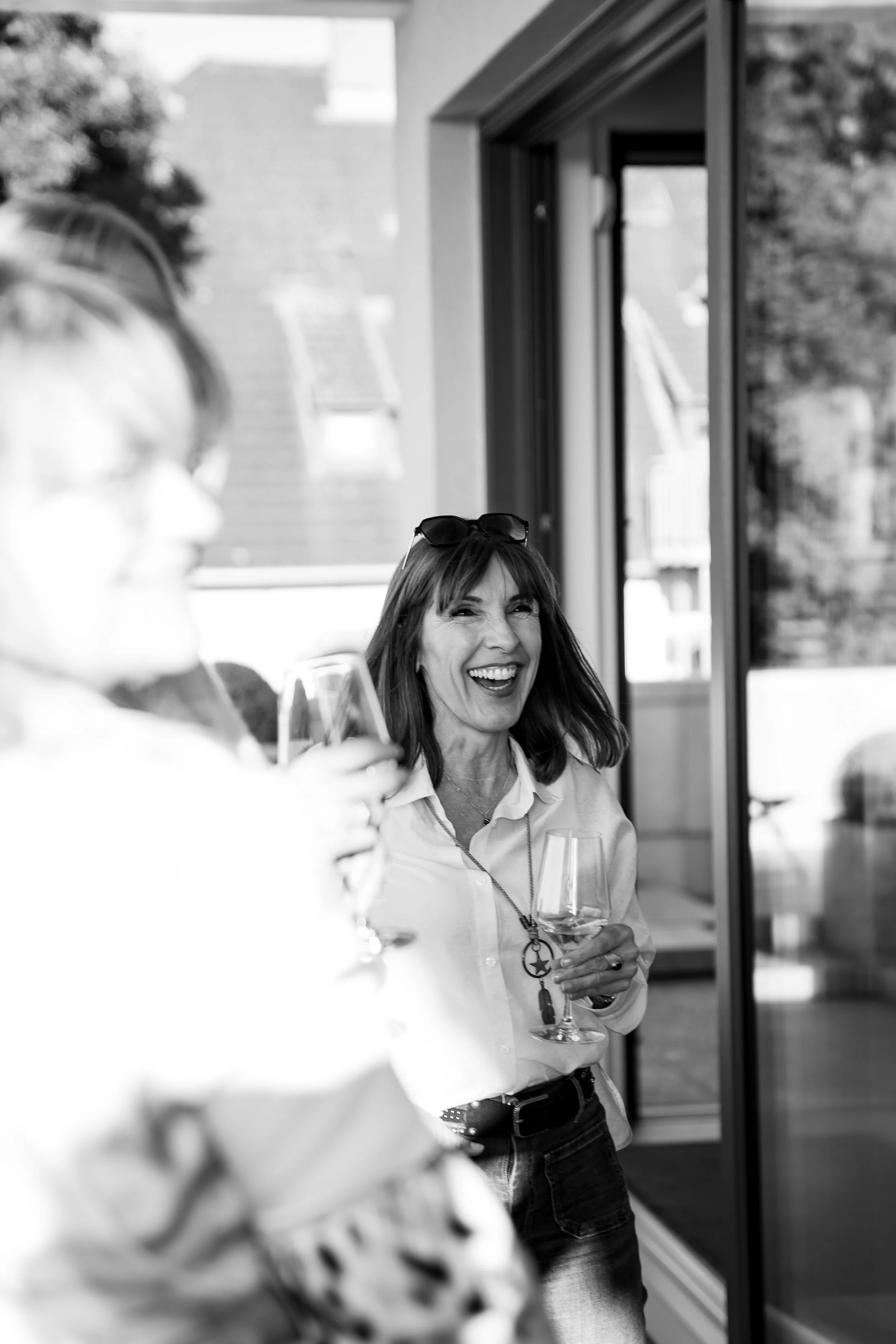 Eine lachende Frau mit dunklen Haaren, Sonnenbrille auf dem Kopf, hält ein Weinglas, im Hintergrund eine offene Tür und eine Frau mit Brille, die ebenfalls ein Weinglas hält, bei einem geselligen Treffen.