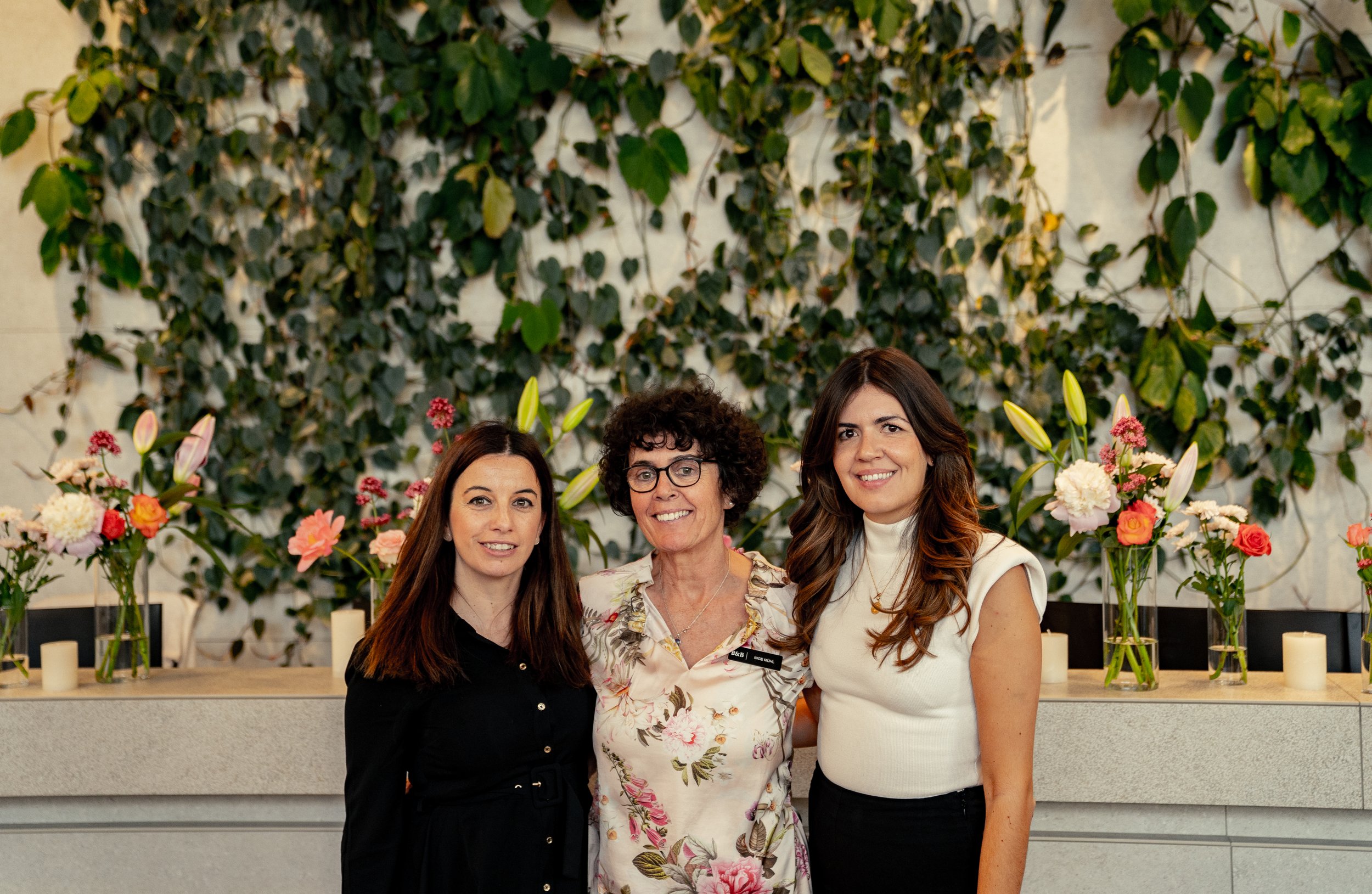 Drei Frauen stehen vor einer mediterranen Pflanzenwand mit Blumen und Kerzen.