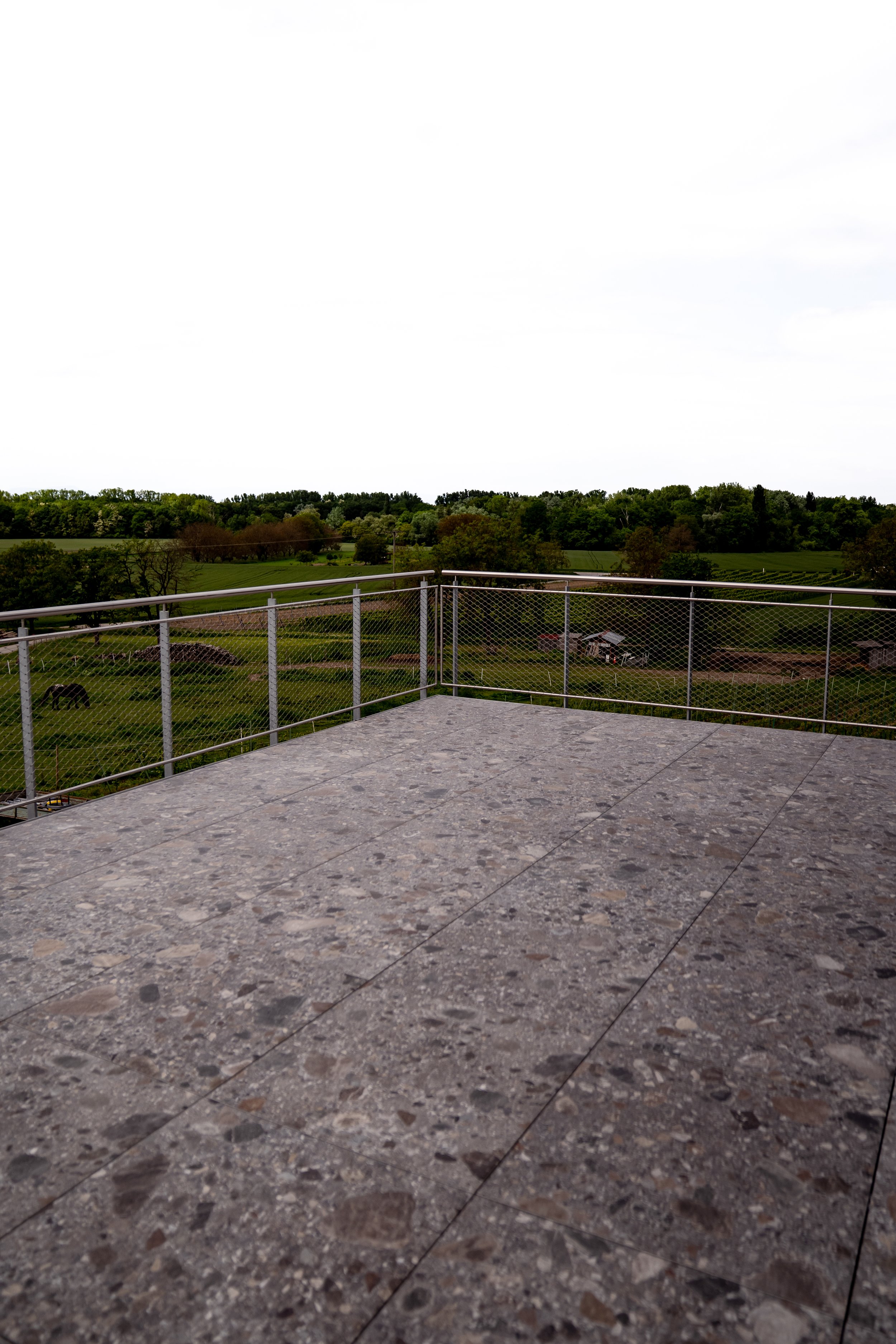 Blick von einer Terrasse mit grauem Boden auf eine grüne Landschaft mit Bäumen und Häusern, umgeben von Feldern, ein Himmel mit Wolken.