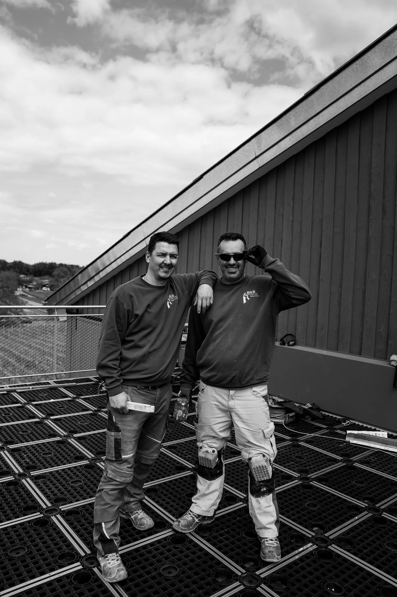 Zwei Männer auf einer Baustelle, die lächelnd in die Kamera schauen, mit einem modernen Gebäude und Wolken im Himmel im Hintergrund.