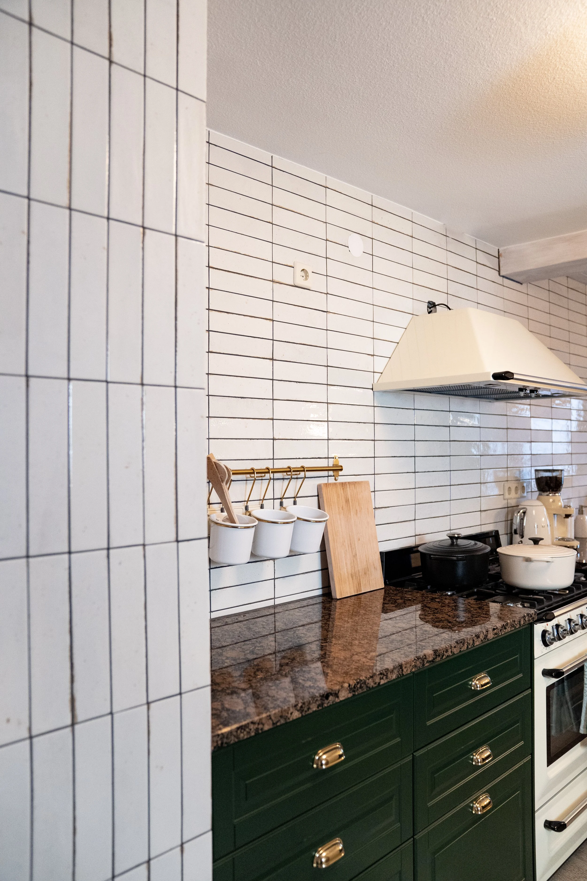 Kücheninsel mit dunklem Granit-Arbeitsplatte, weiße Wandfliesen, grüne Küchenschränke mit goldfarbenen Griffen, Herd mit Kochutensilien, Küchenutensilien an Wand und eine Kaffeemaschine.