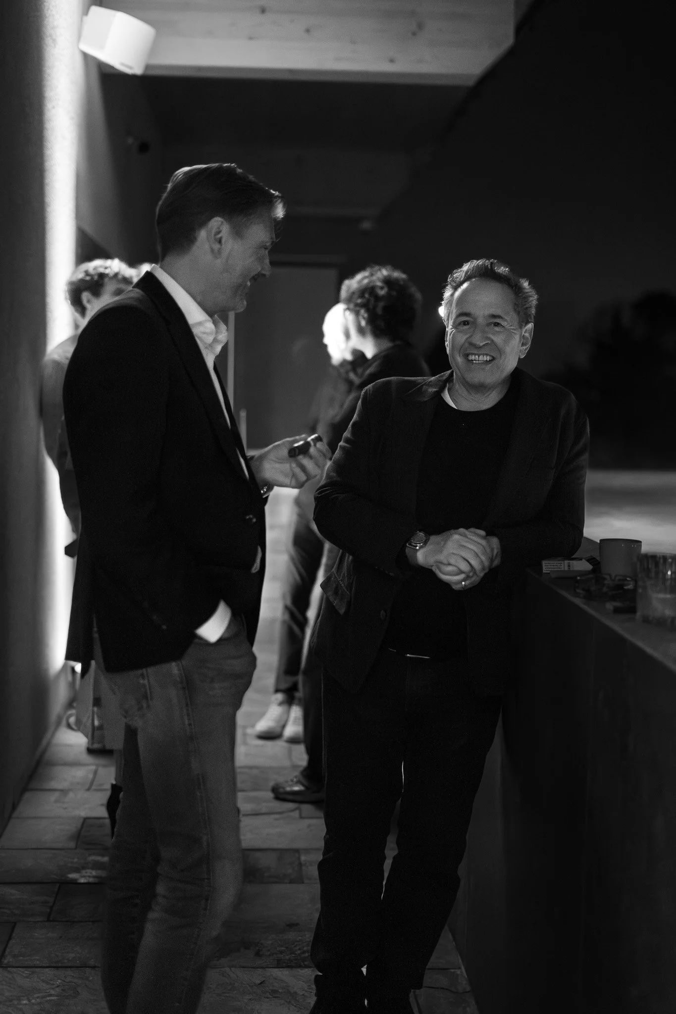 Zwei Männer unterhalten sich bei einer Party, einer lächelt freundlich, die Szenerie ist abends in einem dunklen Raum, im Hintergrund weitere Personen.