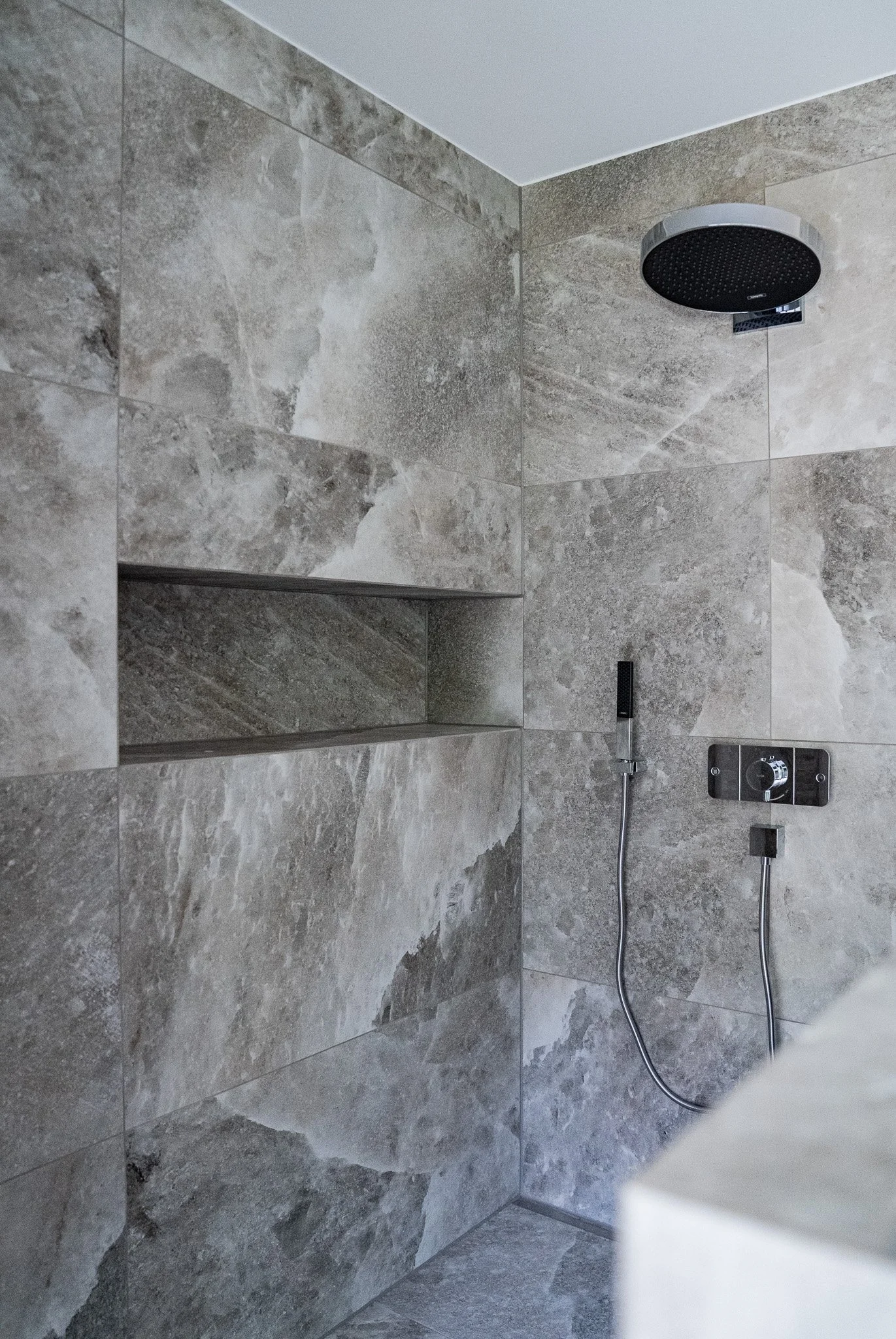 Duschecke mit regulierbarem Duschkopf und Steuerelementen, gefliest mit grauen Natursteinfliesen.