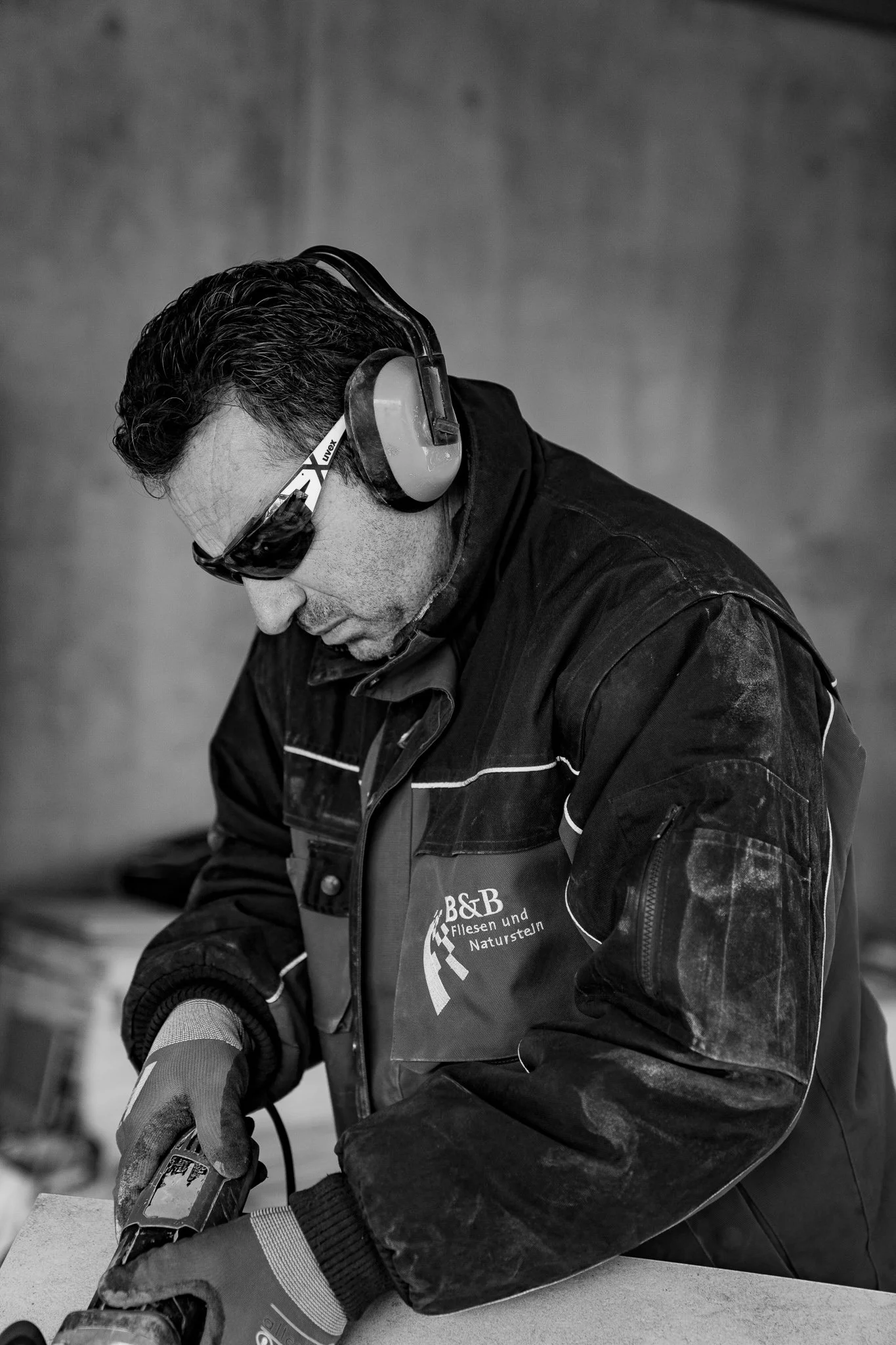 Mann trägt Schutzbrille, Gehörschutz und Arbeitshandschuhe, arbeitet mit einem Werkzeug an einer flachen Oberfläche, im Hintergrund ein ungeordneter Arbeitsplatz, schwarz-weiß Foto.