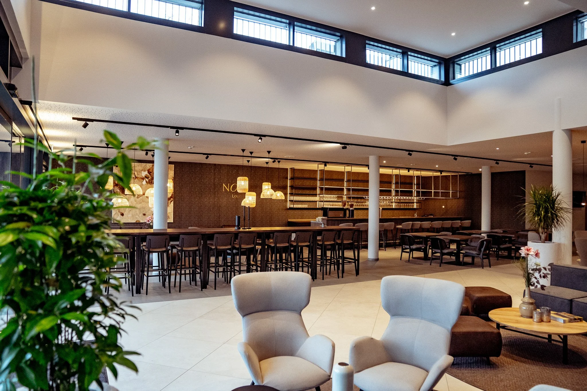 Heller, moderner Lounge mit bequemen Stühlen, einem Couchtisch mit Dekorationen und einem hohen Barbereich im Hintergrund, beleuchtet mit warmem Licht.