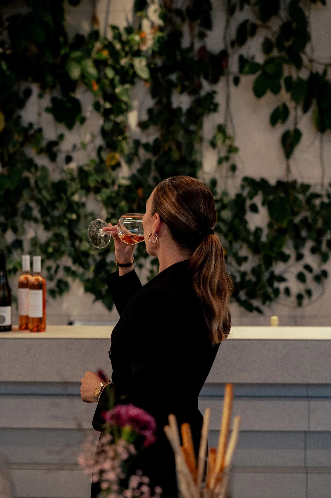 Frau mit braunen Haaren in einem schwarzen Outfit, die ein Glas Roséwein hält, im Hintergrund eine Bar mit Flaschen und eine grüne, bewachsene Wand.