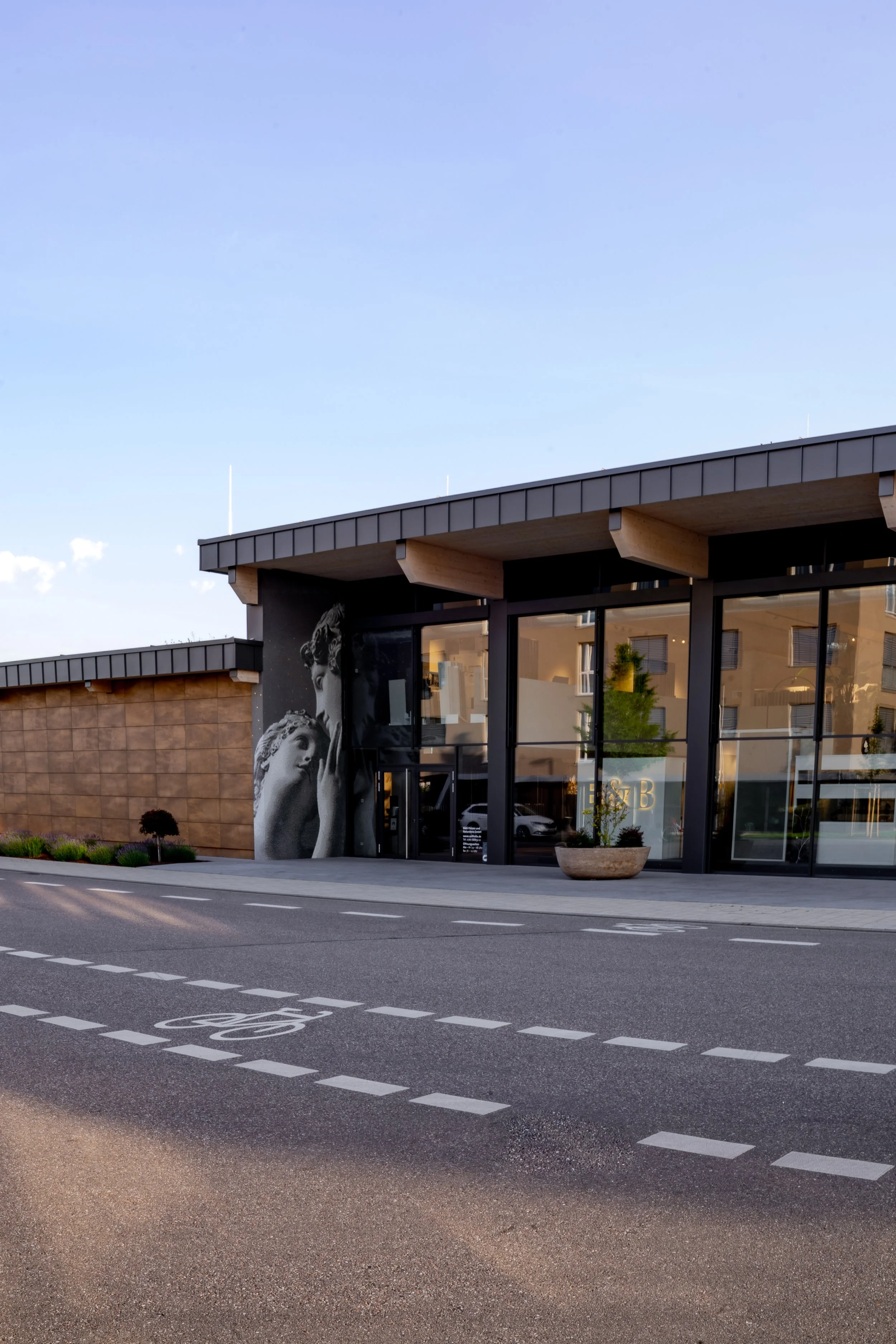 Modernes Gebäude mit großen Glasfenstern und Wandkunst eines Skulpturenduos, umgeben von einem Fahrradweg und einem Parkplatz bei Tageslicht.